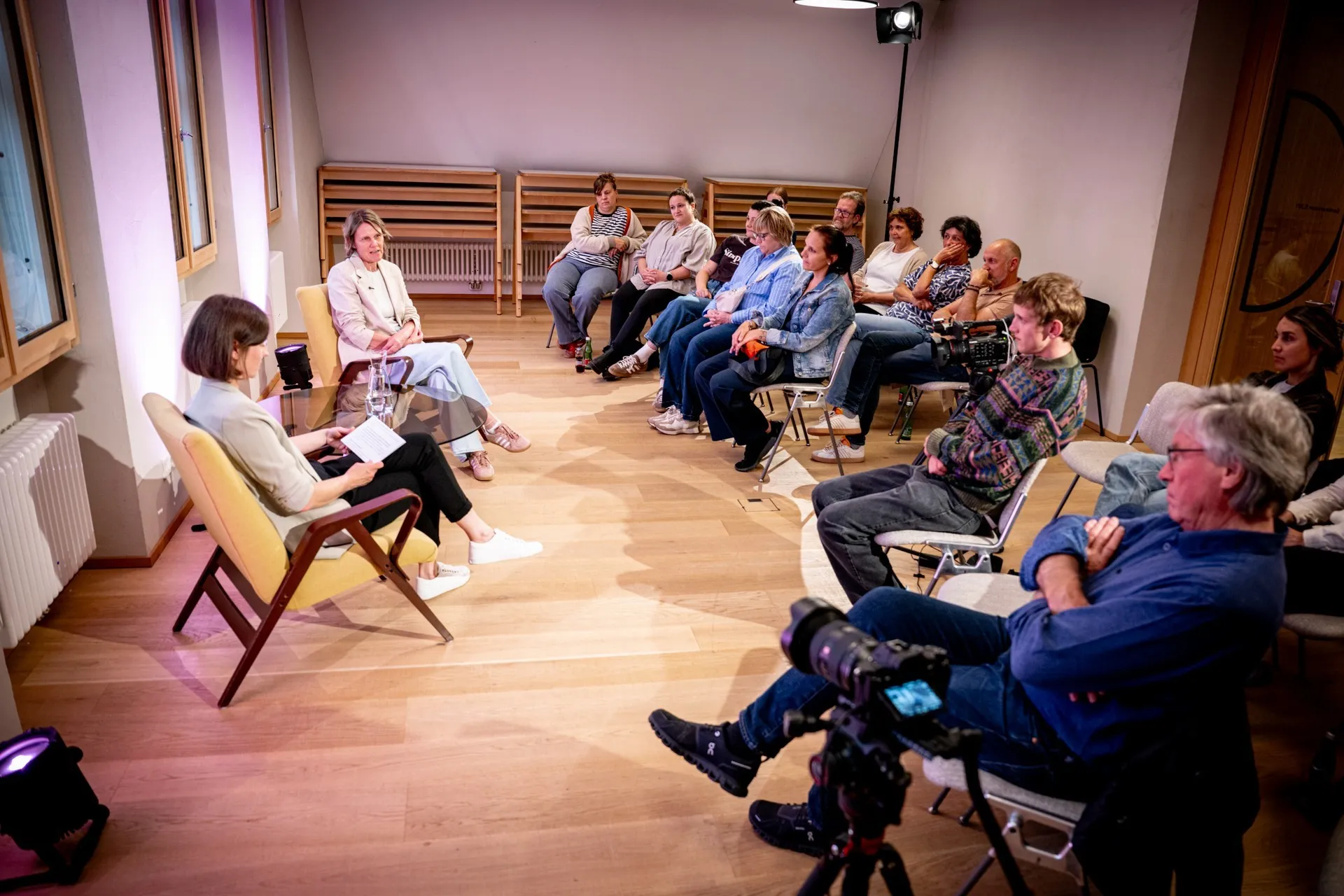 Eine Weitwinkelaufnahme der Podiumsdiskussion aus erhöhter Perspektive. Auf der linken Seite sitzen sich die Moderatorin (Vordergrund, von hinten zu sehen) und Birgit Wrann (Hintergrund) in gelben Sesseln gegenüber. Zwischen ihnen steht ein niedriger Glastisch. Auf der rechten Seite und im Hintergrund sitzt das Publikum im Halbkreis auf Stühlen und lauscht dem Gespräch. Im rechten Vordergrund sitzt ein älterer Mann im blauen Jeanshemd mit verschränkten Armen; vor ihm ist eine professionelle Filmkamera auf einem Stativ positioniert, die auf die beiden Frauen gerichtet ist. Die Wände links werden von Bodenscheinwerfern in rosa-violettes Licht getaucht.