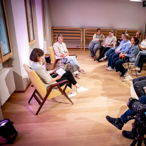 Eine Weitwinkelaufnahme der Podiumsdiskussion aus erhöhter Perspektive. Auf der linken Seite sitzen sich die Moderatorin (Vordergrund, von hinten zu sehen) und Birgit Wrann (Hintergrund) in gelben Sesseln gegenüber. Zwischen ihnen steht ein niedriger Glastisch. Auf der rechten Seite und im Hintergrund sitzt das Publikum im Halbkreis auf Stühlen und lauscht dem Gespräch. Im rechten Vordergrund sitzt ein älterer Mann im blauen Jeanshemd mit verschränkten Armen; vor ihm ist eine professionelle Filmkamera auf einem Stativ positioniert, die auf die beiden Frauen gerichtet ist. Die Wände links werden von Bodenscheinwerfern in rosa-violettes Licht getaucht.