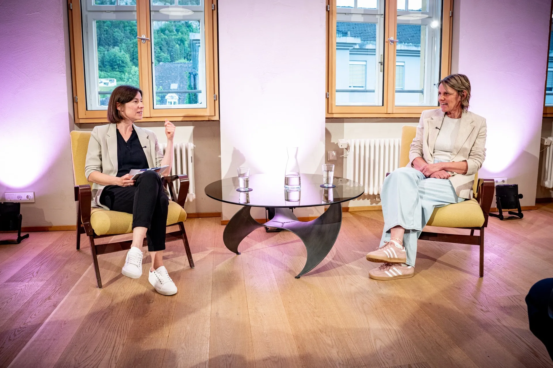 Ein hell erleuchteter Raum mit Holzboden, in dem ein Interview oder eine Podiumsdiskussion stattfindet. Im Hintergrund sitzen zwei Frauen in Sesseln einander gegenüber: Links sitzt eine Moderatorin in dunkler Kleidung und hellem Sakko mit einem Notizblock. Rechts sitzt Birgit Wrann im Rahmen der Veranstaltungsreihe „ArbeitsLebensGeschichte“ der Schaffarei (AK Vorarlberg). Sie trägt ein helles Outfit und spricht engagiert. Zwischen ihnen steht ein niedriger Glastisch mit Wassergläsern und einer Karaffe. Die Szene wird von unten mit rosa-violettem Scheinwerferlicht an den Wänden atmosphärisch beleuchtet. Im Vordergrund sieht man die Rückansicht von Zuschauenden auf Stühlen sowie links eine professionelle Filmkamera auf einem Stativ, die die beiden Frauen aufnimmt.