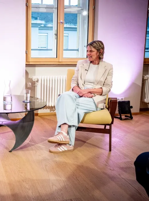Ein hell erleuchteter Raum mit Holzboden, in dem ein Interview oder eine Podiumsdiskussion stattfindet. Im Hintergrund sitzen zwei Frauen in Sesseln einander gegenüber: Links sitzt eine Moderatorin in dunkler Kleidung und hellem Sakko mit einem Notizblock. Rechts sitzt Birgit Wrann im Rahmen der Veranstaltungsreihe „ArbeitsLebensGeschichte“ der Schaffarei (AK Vorarlberg). Sie trägt ein helles Outfit und spricht engagiert. Zwischen ihnen steht ein niedriger Glastisch mit Wassergläsern und einer Karaffe. Die Szene wird von unten mit rosa-violettem Scheinwerferlicht an den Wänden atmosphärisch beleuchtet. Im Vordergrund sieht man die Rückansicht von Zuschauenden auf Stühlen sowie links eine professionelle Filmkamera auf einem Stativ, die die beiden Frauen aufnimmt.