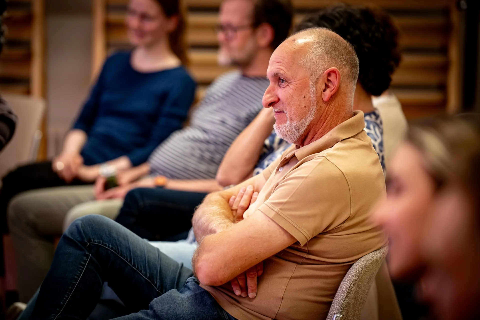 Nahaufnahme eines Zuschauers im Profil während der Veranstaltung. Im Fokus sitzt ein älterer Mann mit Glatze, grauem Vollbart und einem beigefarbenen Poloshirt mit verschränkten Armen. Er blickt mit einem interessierten, leichten Lächeln nach links. Im Hintergrund sind weitere Personen des Publikums unscharf zu sehen, darunter der Mann im gestreiften T-Shirt und die junge Frau im dunkelblauen Oberteil aus den vorherigen Bildern. Im rechten Vordergrund ist das Gesicht einer weiteren Person stark unscharf angeschnitten.