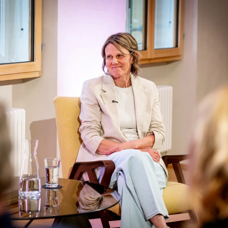 Birgit Wrann sitzt bei einer Gesprächsveranstaltung der ArbeitsLebensGeschichte der Schaffarei der AK Vorarlberg auf einem Sessel und spricht vor Publikum. Sie trägt einen hellen Blazer und lächelt in Richtung der Zuhörenden. Im Vordergrund sind verschwommen Gäste sowie ein Tisch mit Wasserkrug und Glas zu sehen.