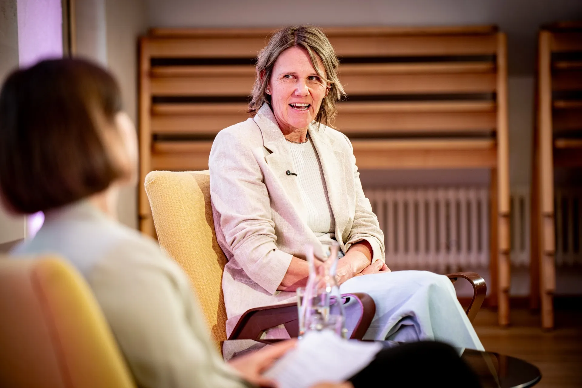 Porträtaufnahme von Birgit Wrann während einer Podiumsdiskussion. Sie sitzt leicht nach links gedreht in einem gelben Sessel, blickt ihre Gesprächspartnerin an und spricht mit lebendigem, lächelndem Gesichtsausdruck. Sie trägt ein helles Sakko über einem weißen Oberteil und eine helle Hose. Im linken Vordergrund ist die Moderatorin unscharf von hinten zu sehen, wie sie das Gespräch führt. Im Hintergrund ist eine hölzerne Wandkonstruktion angedeutet, und auf dem Tisch vor den beiden Frauen ist der obere Teil einer Wasserkaraffe zu erkennen.