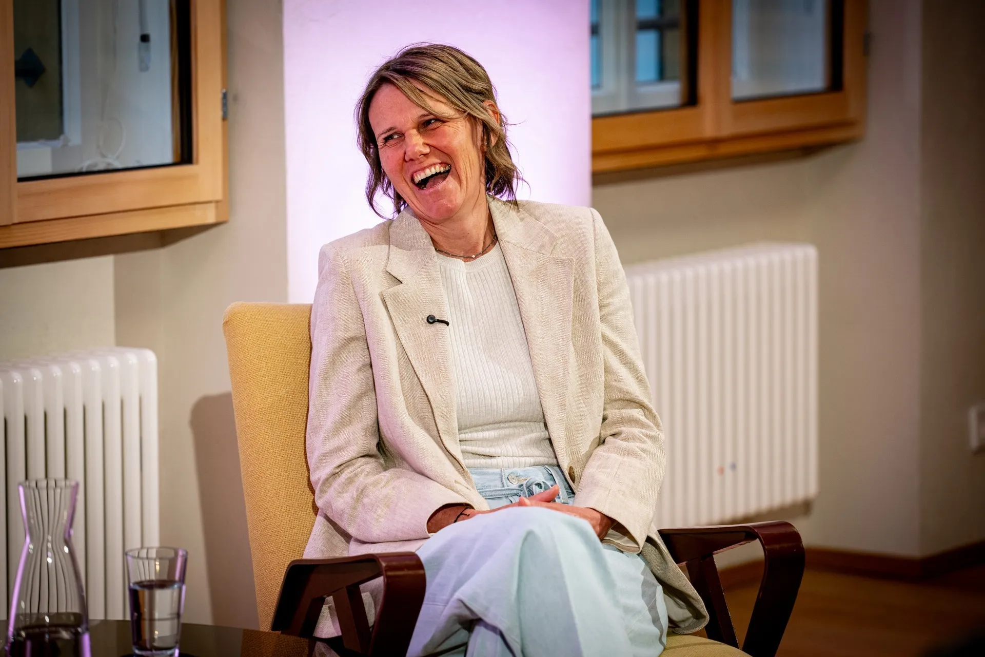Halbporträt von Birgit Wrann während der Podiumsdiskussion. Sie sitzt in einem gelben Sessel, hat die Beine überschlagen und lacht herzlich mit geöffnetem Mund, während sie den Kopf leicht nach links neigt. Sie trägt ein helles, beigefarbenes Sakko über einem feingerippten weißen Oberteil und eine helle Jeans. Am Kragen ihres Sakkos ist ein kleines Ansteckmikrofon befestigt. Im linken Vordergrund sind eine Wasserkaraffe und ein gefülltes Glas auf einem Tisch angeschnitten. Der Hintergrund zeigt die helle Wand des Raumes, ein Holzfenster und einen Heizkörper.