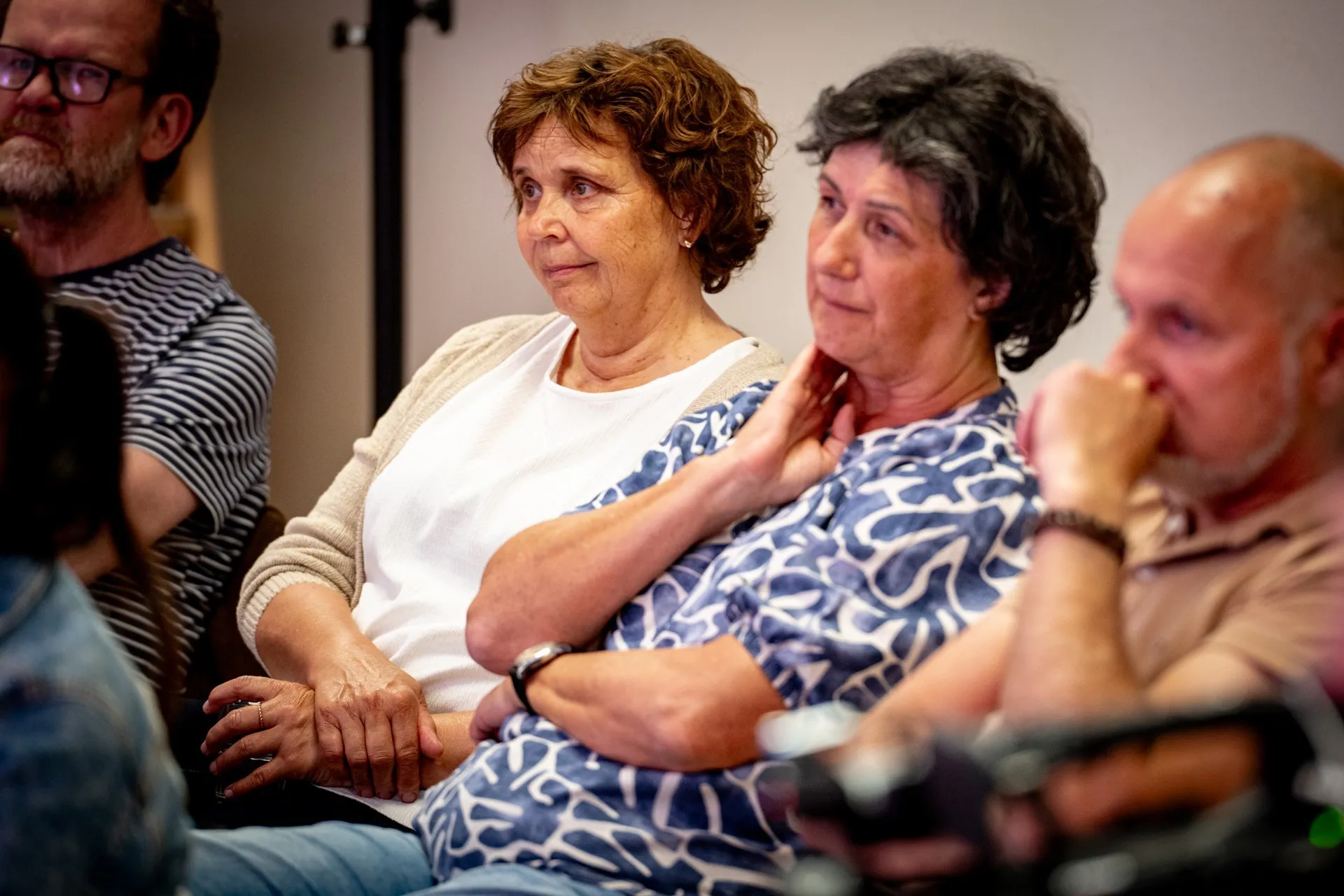 Nahaufnahme von Zuschauenden im Publikum der Veranstaltung. Im Mittelpunkt sitzen zwei ältere Frauen nebeneinander und blicken aufmerksam nach links. Die linke Frau hat braune, lockige Haare und trägt eine helle Strickjacke über einem weißen Oberteil. Die rechte Frau hat dunkle, kurze Haare, trägt eine blau-weiß gemusterte Bluse und hält eine Hand leicht an den Hals. Links im Hintergrund ist der Mann mit Brille und gestreiftem T-Shirt zu sehen. Rechts im Vordergrund sitzt unscharf der Mann im beigefarbenen Poloshirt mit nachdenklich an den Mund gelegter Hand.