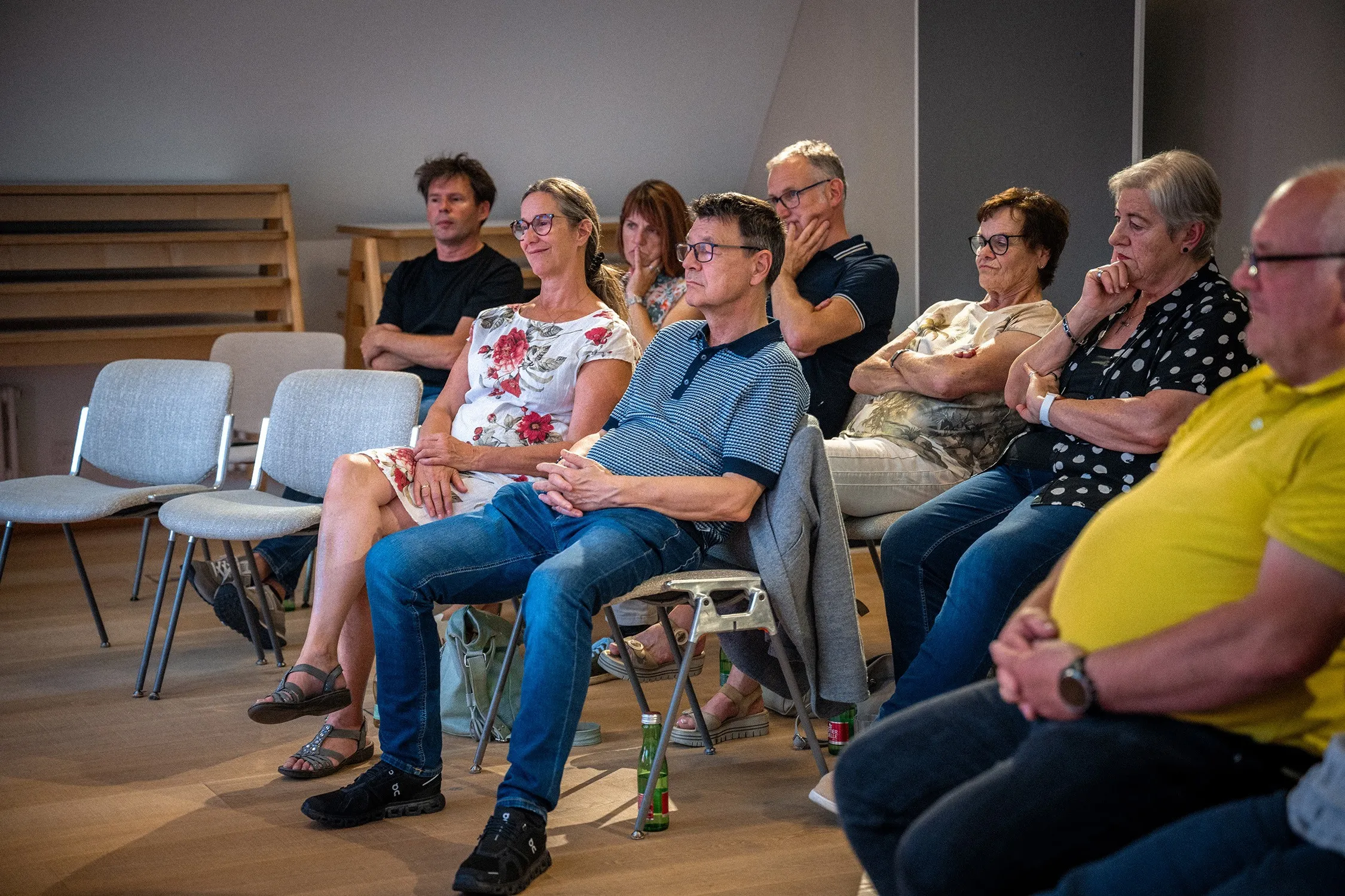 Eine Reihe von Zuschauerinnen und Zuschauern sitzt auf hellen Stühlen in einem modernen Raum mit Holzboden und hört fokussiert einem Vortrag zu. Im Zentrum steht ein Mann mit Brille, kurzärmeligem blau gestreiftem Poloshirt und Jeans. Links neben ihm sitzt eine Frau in einem weißen Sommerkleid mit rotem Blumenmuster. Weitere Personen im Hintergrund und an den Seiten folgen der Präsentation aufmerksam.