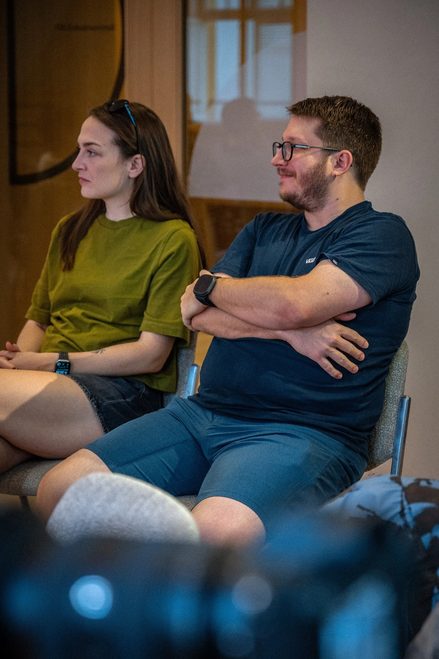 Eine Frau und ein Mann sitzen nebeneinander im Publikum. Rechts sitzt ein Mann mit kurzer Barttracht, Brille, dunklem T-Shirt und blauen Shorts, der mit verschränkten Armen lächelnd zur Seite blickt. Links von ihm sitzt eine Frau mit langen braunen Haaren, einer Sonnenbrille auf dem Kopf, einem olivenen T-Shirt und kurzen dunklen Hosen. Beide tragen eine Smartwatch am Handgelenk und blicken aufmerksam in dieselbe Richtung.