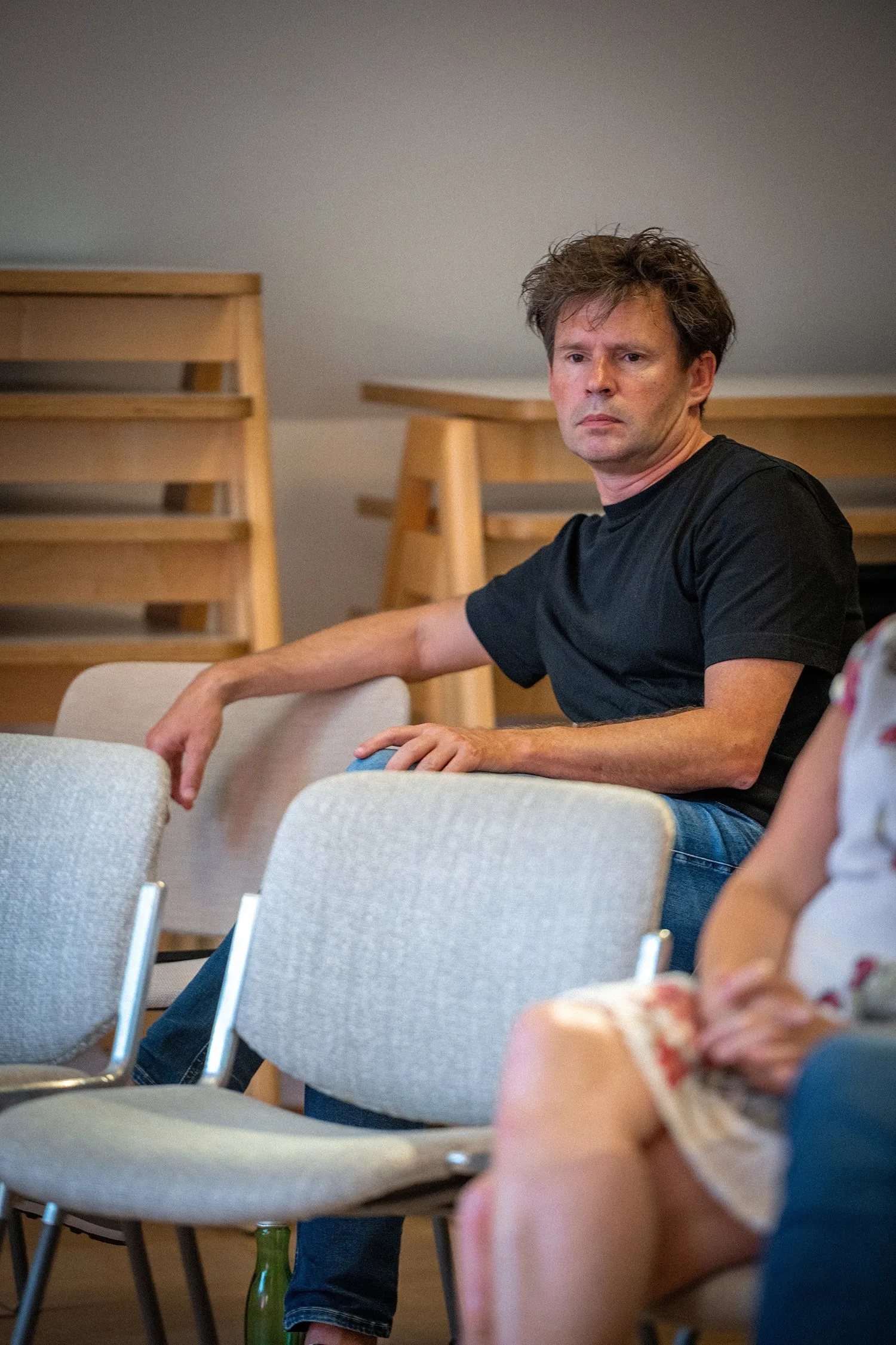 Ein Mann mittleren Alters mit wuscheligem dunklem Haar, schwarzem T-Shirt und Jeans sitzt im Publikum. Er stützt einen Arm auf die Stuhllehne neben sich und schaut mit einem ernsten, aufmerksamen Blick leicht nach rechts. Im unscharfen Vordergrund sieht man weitere Stühle und eine Person, im Hintergrund stehen gestapelte Holzmöbel.
