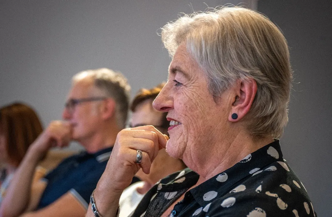 Eine Frau mit kurzem, ergrautem Haar sitzt im Profil im Vordergrund und schaut mit einem leichten Lächeln nach links. Sie trägt eine kurzärmelige, schwarz-weiß gepunktete Bluse, eine helle Smartwatch am Handgelenk sowie Ringe und ein Armband. Eine Hand stützt sie sanft unter dem Kinn ab. Im unscharfen Hintergrund sitzen weitere Personen, darunter ein Mann mit Brille, die ebenfalls aufmerksam in dieselbe Richtung blicken.