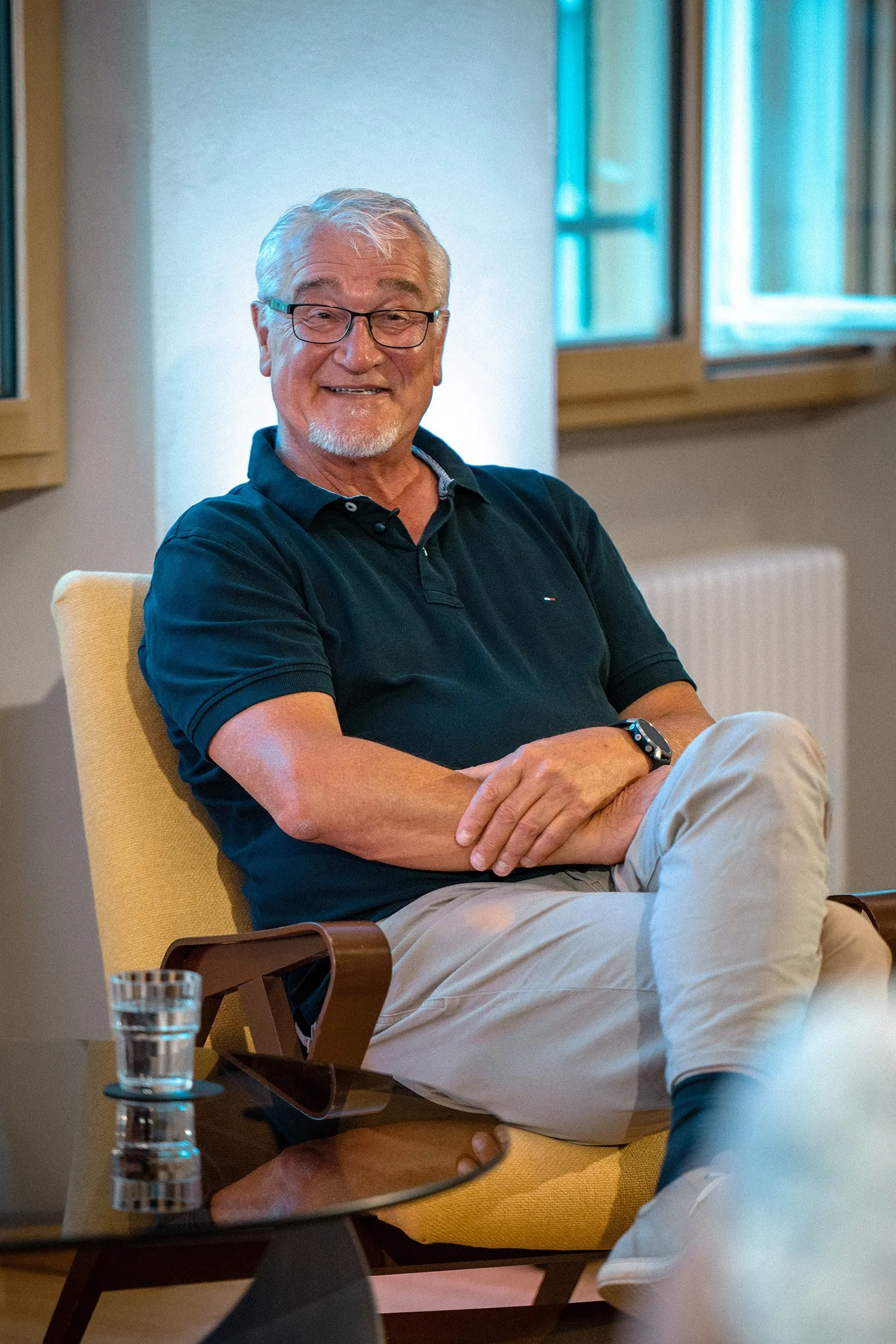 Porträt von Mike Reisinger, einem älteren Mann mit kurzen grauen Haaren, einer rechteckigen Brille und einem Goatee-Bart. Er sitzt entspannt in einem gelben Sessel, hat die Beine übereinandergeschlagen und die Arme vor der Brust verschränkt. Er trägt ein dunkelblaues Poloshirt, eine helle Hose und eine dunkle Uhr am Handgelenk. Auf dem Glastisch vor ihm steht ein Wasserglas. Im Hintergrund ist ein hell erleuchteter Raum mit Holzfensterrahmen zu sehen.