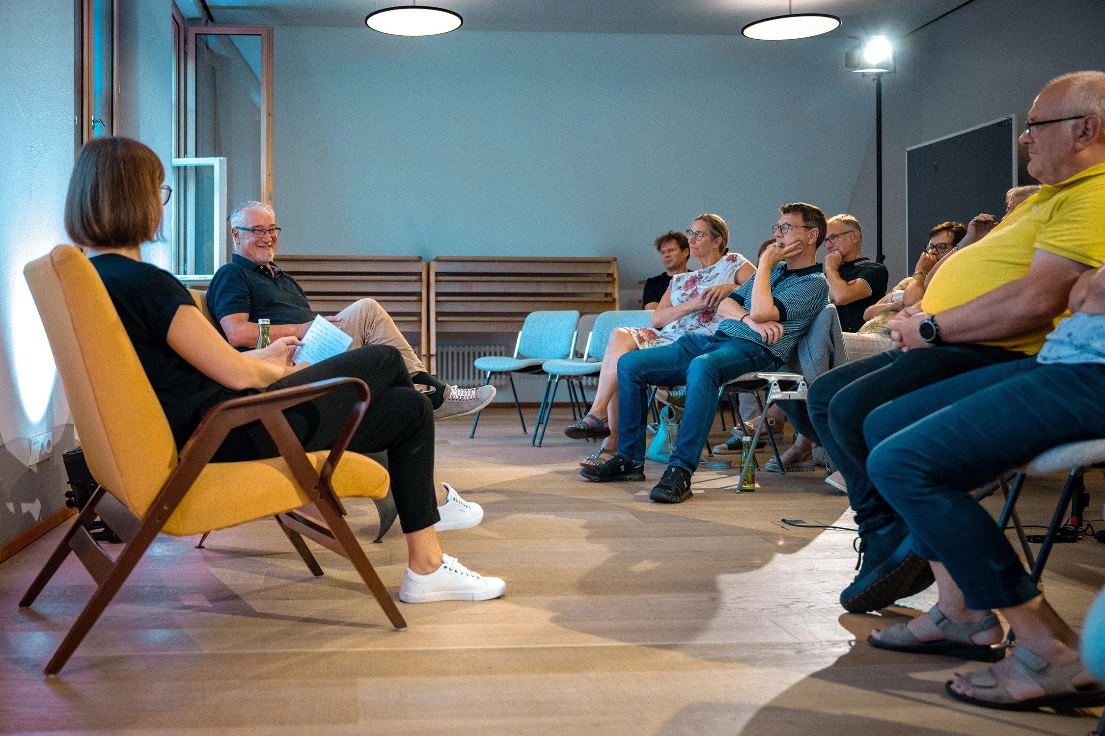 Gesamtaufnahme einer Podiumsdiskussion in einem modernen Raum mit Holzboden und runden Deckenleuchten. Links sitzt die Moderatorin im gelben Sessel mit dem Rücken teilweise zur Kamera und hält Notizen. Ihr gegenüber sitzt Mike Reisinger, ein ergrauter Mann mit Brille, der sie lächelnd anblickt. Rechts sitzt das Publikum auf Stühlen in U-Form angeordnet und verfolgt das Gespräch aufmerksam.