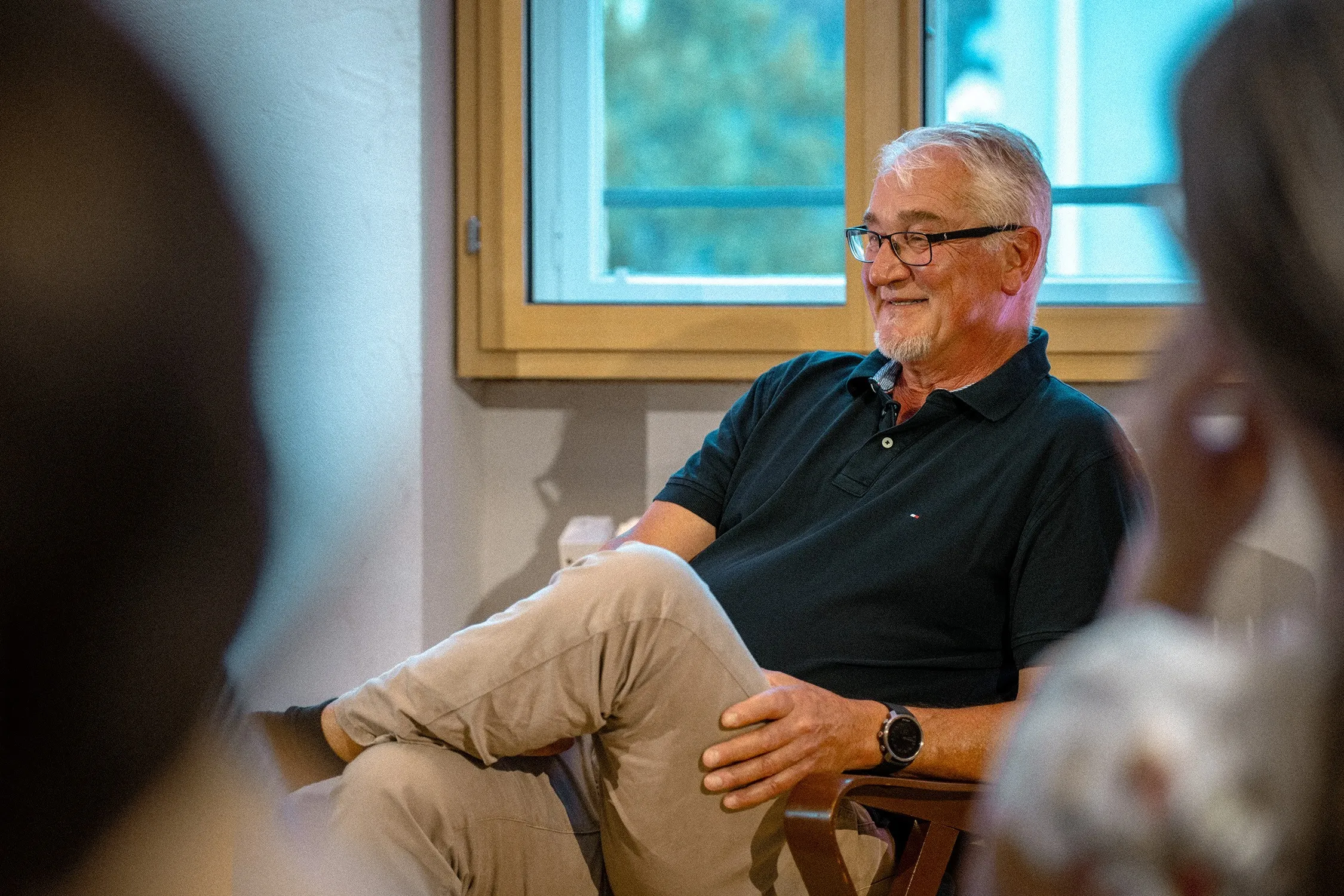 Mike Reisinger, ein älterer Mann mit kurzen grauen Haaren, Goatee-Bart und Brille sitzt entspannt in einem Sessel. Er trägt ein dunkelblaues Poloshirt sowie eine helle Hose, hat die Beine übereinandergeschlagen und stützt eine Hand auf dem Knie ab. Er schaut mit einem freundlichen Lächeln leicht zur Seite. Im Hintergrund ist ein großes Fenster mit Holzrahmen zu sehen, während im Vordergrund unscharf die Silhouetten anderer Personen zu erkennen sind.
