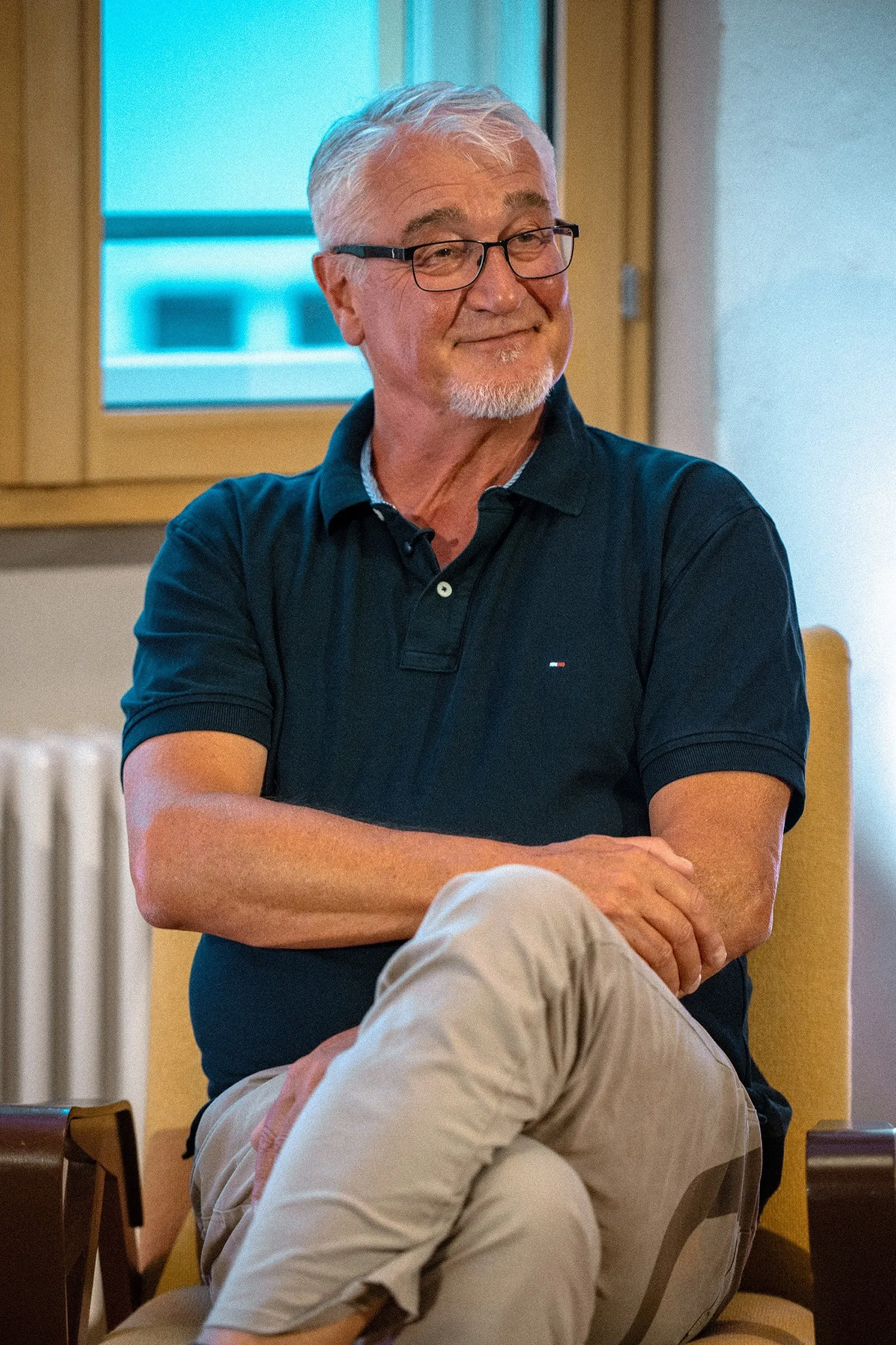 Nahaufnahme von Mike Reisinger, einesm älteren Mann mit kurzen grauen Haaren, Goatee-Bart und Brille mit dunklem Gestell. Er sitzt im gelben Sessel, hat die Beine übereinandergeschlagen und blickt mit einem nachdenklichen, sanften Lächeln leicht nach rechts. Er trägt ein dunkelblaues Poloshirt und eine helle Hose. Im Hintergrund ist ein Fensterrahmen aus Holz zu sehen.