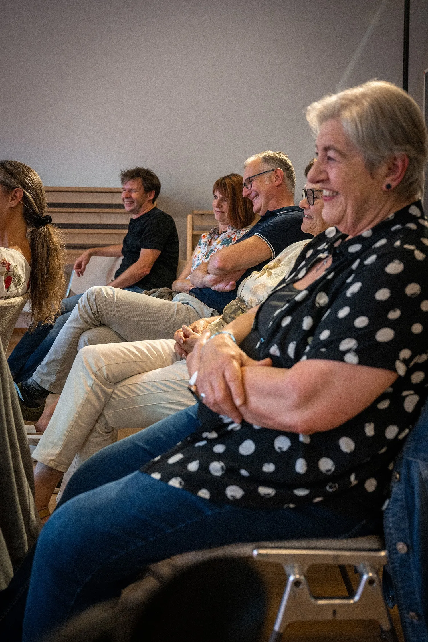 Eine Gruppe von Menschen sitzt nebeneinander in Stuhlreihen bei einer Veranstaltung und schaut lächelnd und aufmerksam nach rechts außen. Im unscharfen Vordergrund befindet sich eine ältere Frau mit grauen, kurzen Haaren, die eine kurzärmelige, schwarz-weiß gepunktete Bluse und Jeans trägt. Neben ihr sitzen weitere Frauen und Männer unterschiedlichen Alters im Profil, die entspannt zuhören, lachen und verschränkte Arme haben. Der Raum ist hell und schlicht gehalten.