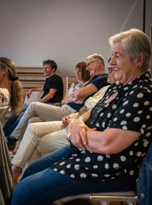 Eine Gruppe von Menschen sitzt nebeneinander in Stuhlreihen bei einer Veranstaltung und schaut lächelnd und aufmerksam nach rechts außen. Im unscharfen Vordergrund befindet sich eine ältere Frau mit grauen, kurzen Haaren, die eine kurzärmelige, schwarz-weiß gepunktete Bluse und Jeans trägt. Neben ihr sitzen weitere Frauen und Männer unterschiedlichen Alters im Profil, die entspannt zuhören, lachen und verschränkte Arme haben. Der Raum ist hell und schlicht gehalten.