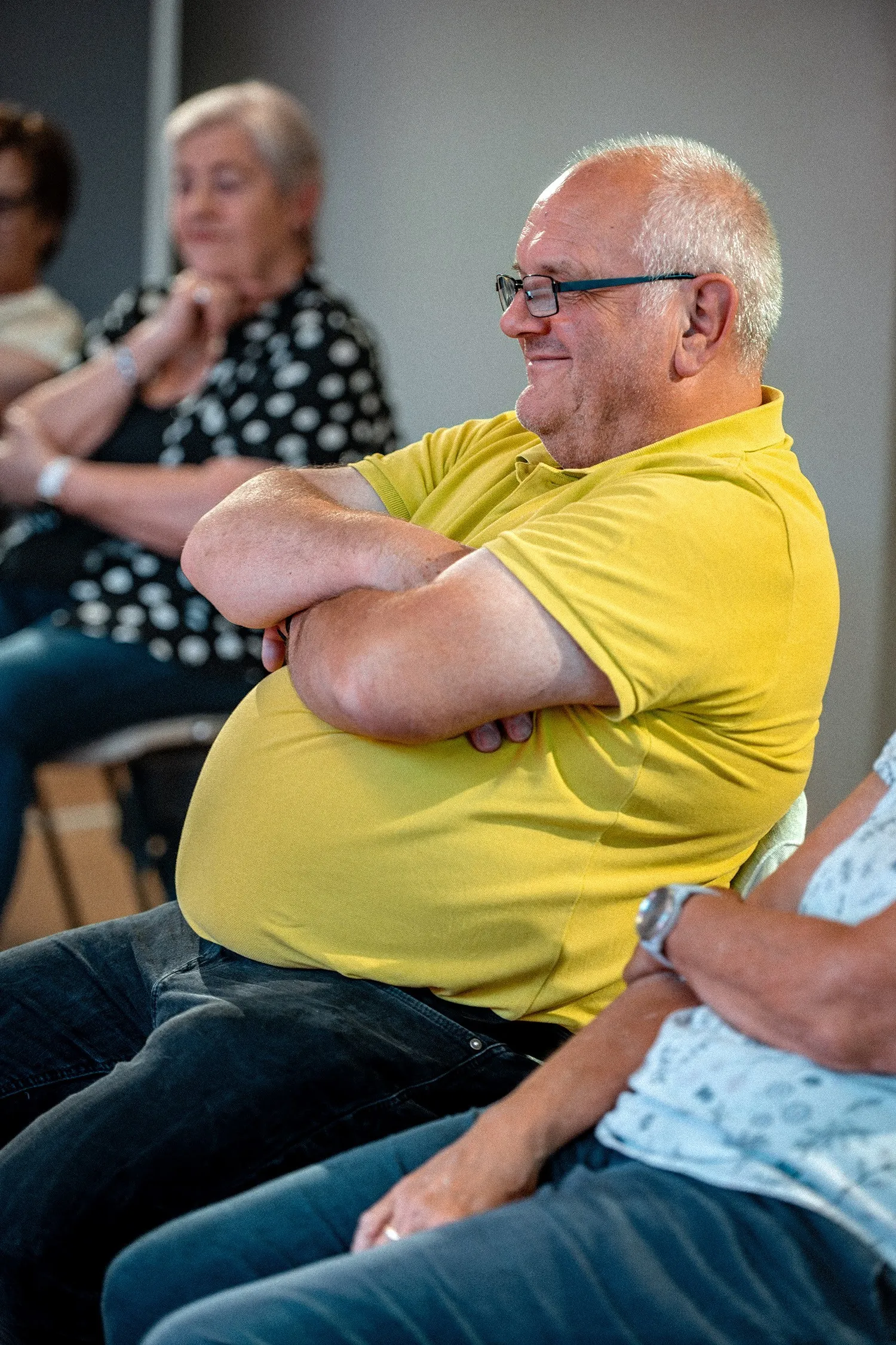 Porträt eines älteren Mannes im Profil, der entspannt im Publikum sitzt. Er trägt eine Sehhilfe mit dunklem Gestell, ein knallgelbes Poloshirt und eine dunkle Hose. Er hat die Arme gemütlich über dem Bauch verschränkt und blickt mit einem zufriedenen Lächeln nach links. Im verschwommenen Hintergrund ist eine Frau mit kurz gelocktem, grauem Haar und einer gepunkteten Bluse zu sehen.