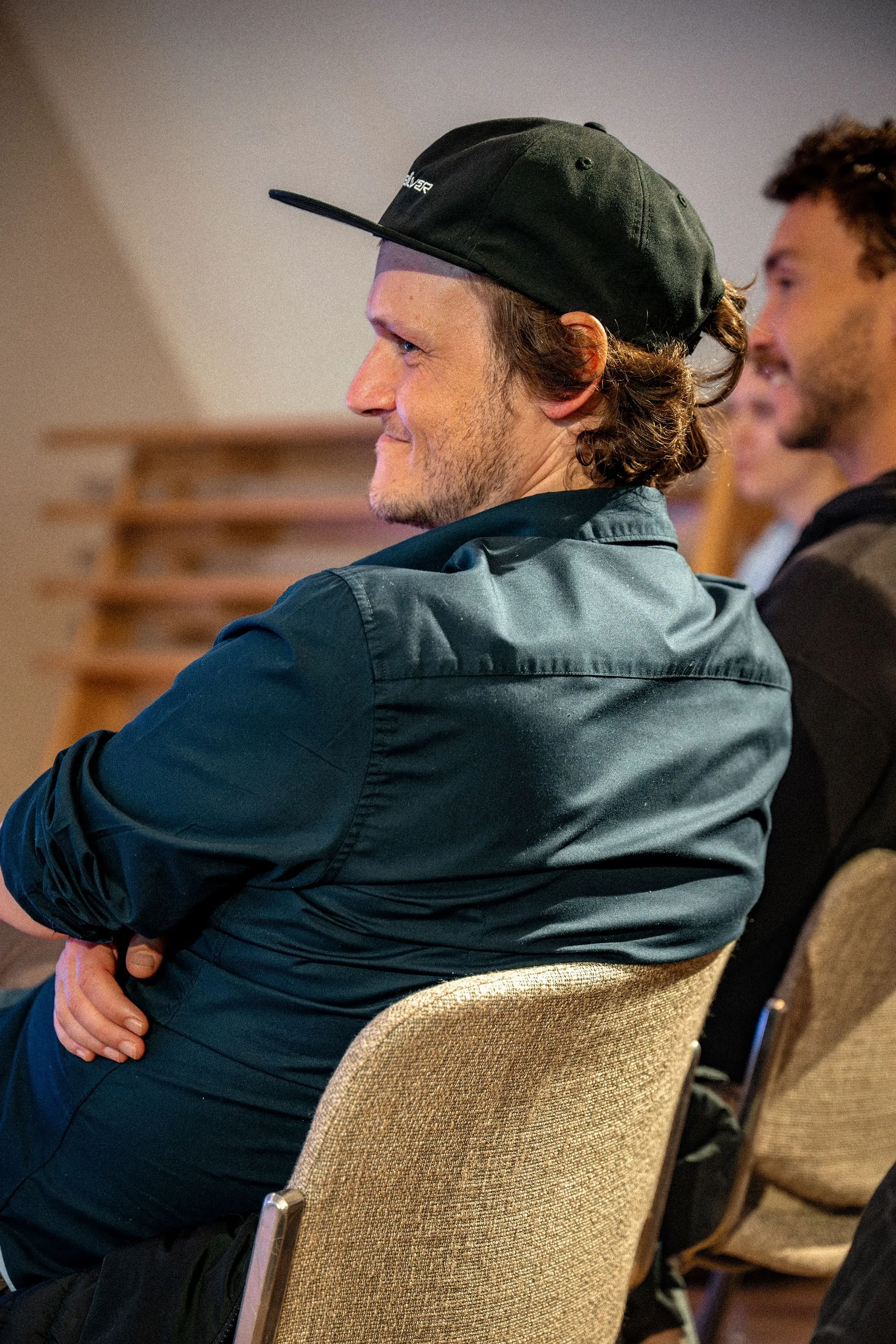 Ein junger Mann sitzt im Publikum und lächelt, leicht zur Seite gedreht. Er trägt ein dunkelblau-grünes Hemd und eine schwarze Snapback-Cap, sein lockiges Haar ist im Nacken zusammengebunden. Im Hintergrund ist ein weiterer Zuhörer zu erkennen. Die Aufnahme entstand bei der Veranstaltung „ArbeitsLebensGeschichten" in der Schaffarei der Arbeiterkammer Vorarlberg in Feldkirch.