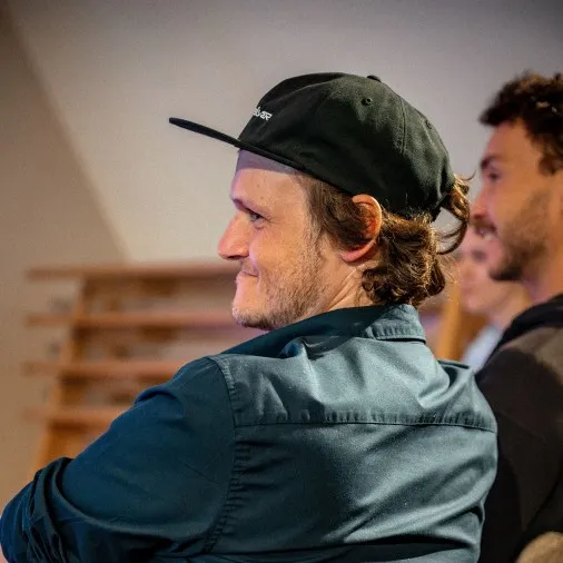 Ein junger Mann sitzt im Publikum und lächelt, leicht zur Seite gedreht. Er trägt ein dunkelblau-grünes Hemd und eine schwarze Snapback-Cap, sein lockiges Haar ist im Nacken zusammengebunden. Im Hintergrund ist ein weiterer Zuhörer zu erkennen. Die Aufnahme entstand bei der Veranstaltung „ArbeitsLebensGeschichten" in der Schaffarei der Arbeiterkammer Vorarlberg in Feldkirch.