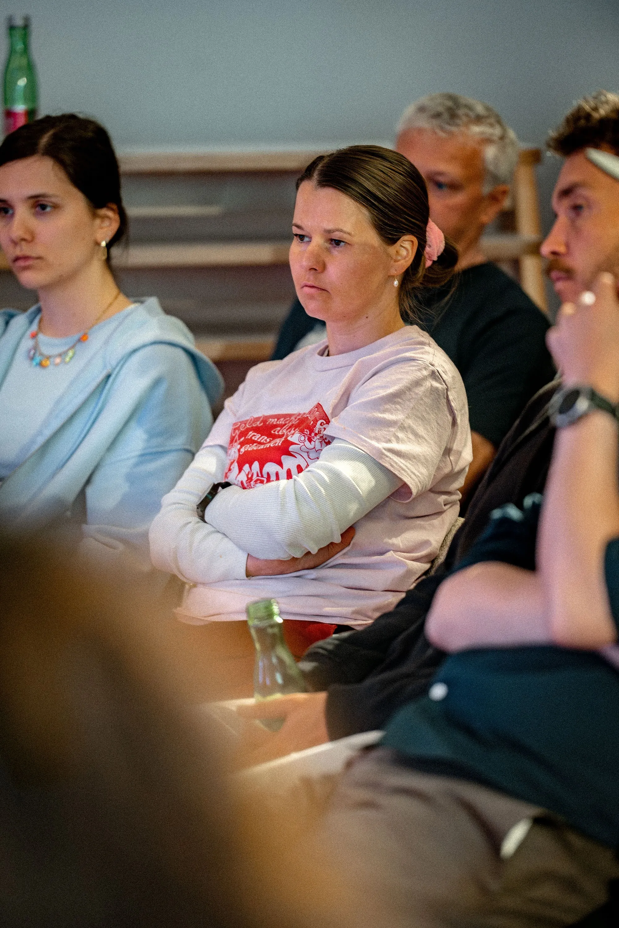 Mehrere Personen sitzen konzentriert im Publikum der Veranstaltung „ArbeitsLebensGeschichten" in der Schaffarei der Arbeiterkammer Vorarlberg in Feldkirch. Im Vordergrund eine Frau mit zurückgebundenem dunklen Haar und Perlenohrringen, die die Arme verschränkt hat und aufmerksam zuhört. Sie trägt ein weißes Langarmshirt unter einem hellrosa T-Shirt mit rotem Aufdruck. Links neben ihr eine jüngere Frau mit dunklem Haar in einem hellblauen Hoodie und bunter Halskette. Im Hintergrund sind weitere Zuschauende sowie hölzerne Sitzstufen zu erkennen.