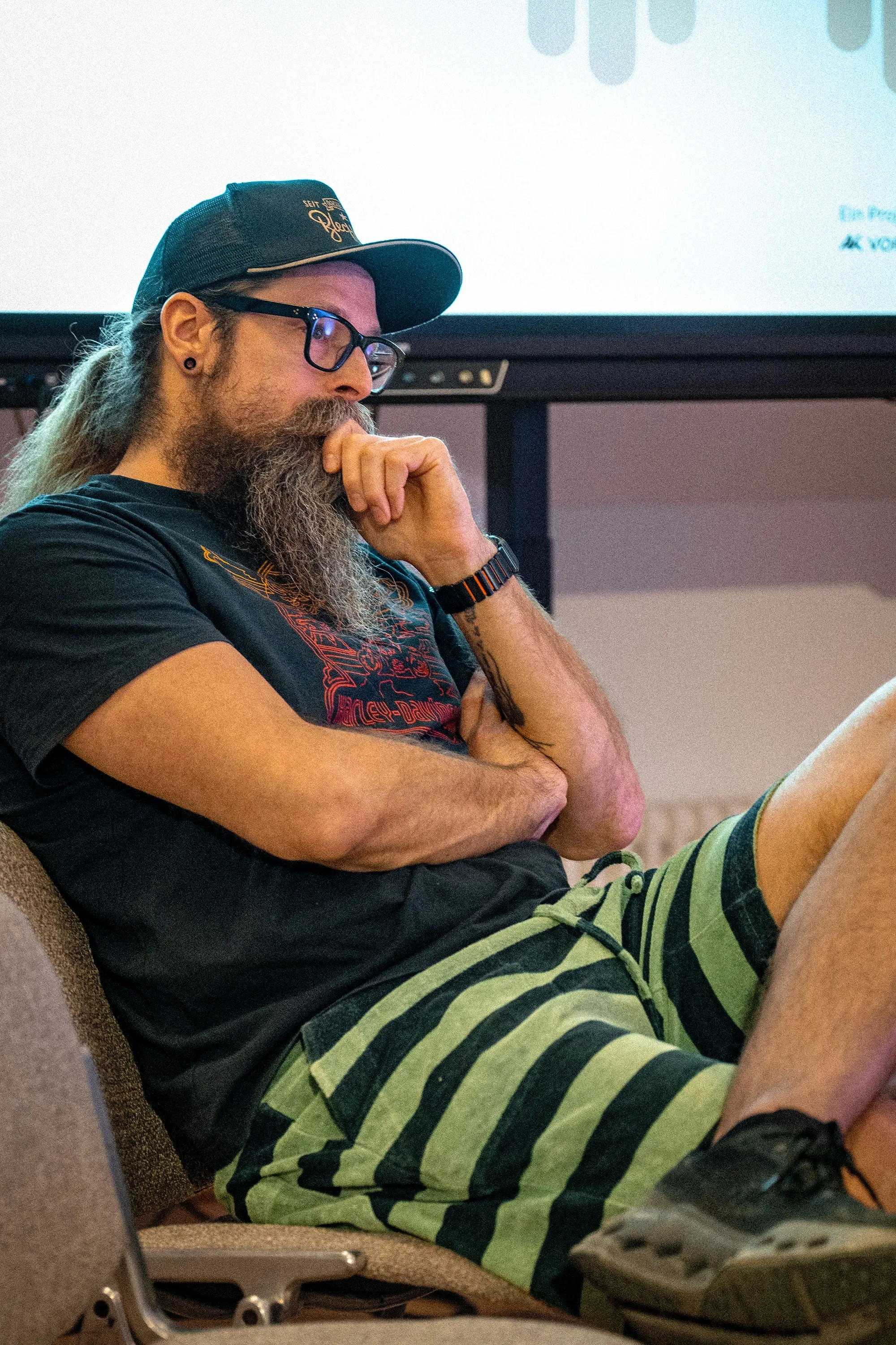 Ein Mann sitzt entspannt und aufmerksam zugehörend auf einem Stuhl. Er trägt ein schwarzes Harley-Davidson-T-Shirt, eine grün-schwarz gestreifte Bermudashorts, eine schwarze Brille und eine dunkle Truckerkappe. Sein langer, grau-brauner Bart fällt auf die Brust, die Hand stützt nachdenklich das Kinn. Im Hintergrund ist eine Leinwand mit dem Logo der AK Vorarlberg zu sehen. Das Foto entstand bei der Veranstaltung „ArbeitsLebensGeschichten" in der Schaffarei der Arbeiterkammer Vorarlberg in Feldkirch, bei der Philipp Böhler (MC Pille One) auftrat.