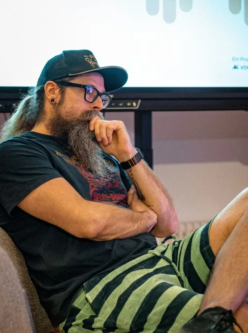 Ein Mann sitzt entspannt und aufmerksam zugehörend auf einem Stuhl. Er trägt ein schwarzes Harley-Davidson-T-Shirt, eine grün-schwarz gestreifte Bermudashorts, eine schwarze Brille und eine dunkle Truckerkappe. Sein langer, grau-brauner Bart fällt auf die Brust, die Hand stützt nachdenklich das Kinn. Im Hintergrund ist eine Leinwand mit dem Logo der AK Vorarlberg zu sehen. Das Foto entstand bei der Veranstaltung „ArbeitsLebensGeschichten" in der Schaffarei der Arbeiterkammer Vorarlberg in Feldkirch, bei der Philipp Böhler (MC Pille One) auftrat.