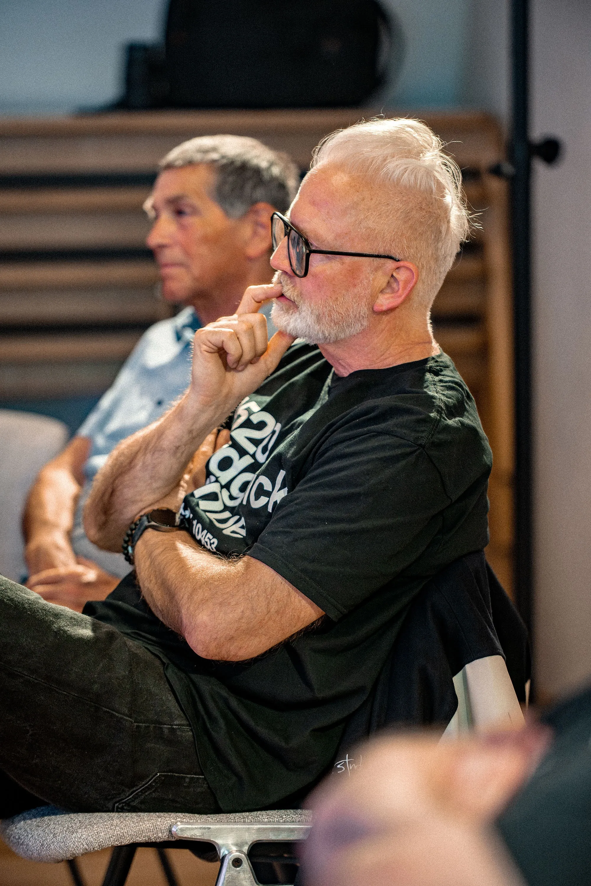 Ein älterer Mann mit weißem Haar und kurzem weißen Bart sitzt nachdenklich im Publikum, die Hand ans Kinn gestützt. Er trägt eine schwarze Brille, ein schwarzes T-Shirt mit weißem Schriftzug sowie eine Perlenkette und eine Uhr am Handgelenk. Im Hintergrund ist ein weiterer Zuhörer mit grauem Haar und hellblauem Hemd zu erkennen, dahinter hölzerne Sitzstufen. Die Aufnahme entstand bei der Veranstaltung „ArbeitsLebensGeschichten" in der Schaffarei der Arbeiterkammer Vorarlberg in Feldkirch.