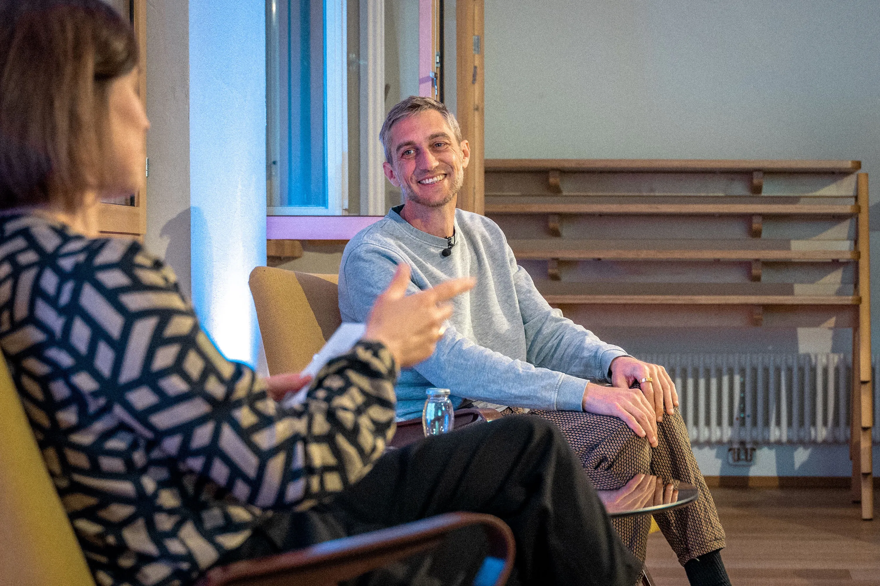 Philipp Böhler (MC Pille One) sitzt bei einer Veranstaltung der ArbeitsLebensGeschichten in der Schaffarei der AK Vorarlberg in Feldkirch auf einem Stuhl und spricht mit einer Moderatorin. Beide sind einander zugewandt, zwischen ihnen steht ein kleiner Tisch mit Wasserflaschen. Die Atmosphäre wirkt ruhig und aufmerksam, der Raum ist modern und warm beleuchtet.