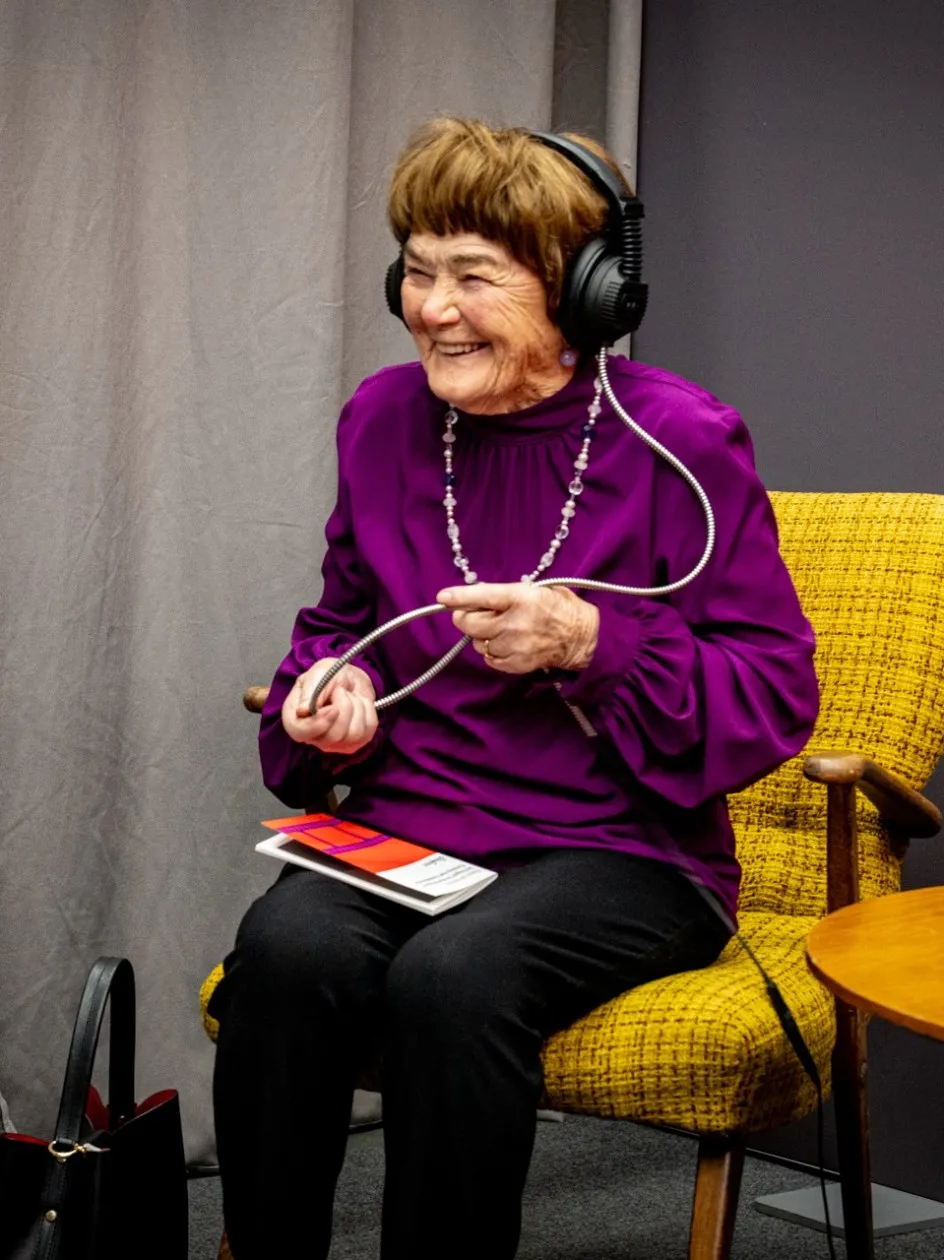 Eine Besucherin der Ausstellung „Museum des Wandels" der AK Vorarlberg sitzt lächelnd in einem gelben Sessel und hört mit einem Kopfhörer ein Audio- oder Video-Interview. In der Hand hält sie das Kabel des Kopfhörers sowie einen Flyer der Ausstellung. Auf dem kleinen Holztisch neben ihr liegen weitere Ausstellungsflyer.