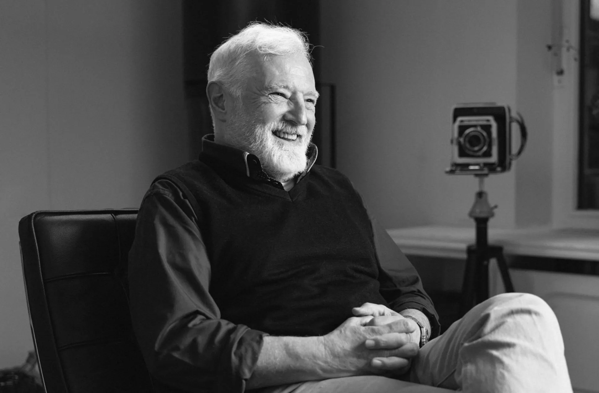 Schwarz-Weiß-Porträt des Fotografen Christian Stuppner: Ein älterer Mann mit weißen Haaren und Bart sitzt entspannt auf einem Stuhl, lächelt und blickt zur Seite. Im Hintergrund steht eine Kamera auf einem Stativ neben einem Fenster und einer Vase.