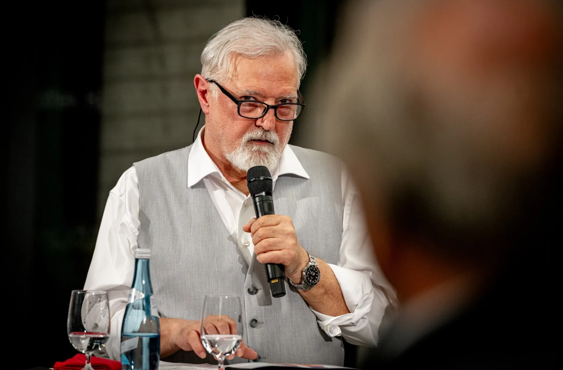 Der Fotograf Christian Stuppner spricht bei der Vernissage zur Ausstellung „Museum des Wandels" der AK Vorarlberg in ein Mikrofon. Er trägt ein weißes Hemd mit grauer Weste und eine schwarze Brille. Vor ihm stehen Gläser und eine Wasserflasche auf dem Tisch.