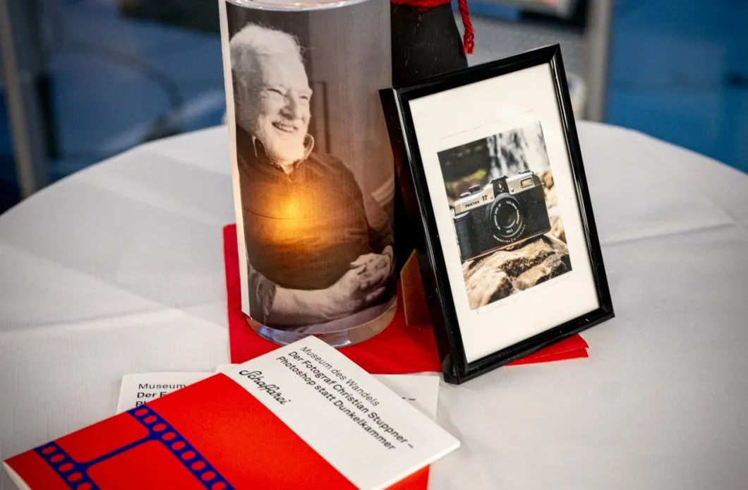 Tischdekoration am Rand der Vernissage zur Ausstellung „Museum des Wandels" der AK Vorarlberg: Programmhefte, ein gerahmtes Bild einer Fotokamera und eine Kerze mit dem Bild des Fotografen Christian Stuppner.