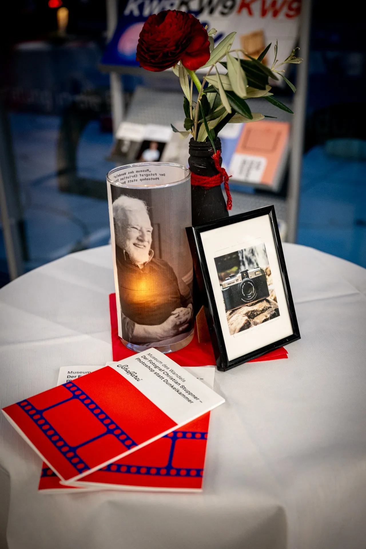 Tischdekoration am Rand der Vernissage zur Ausstellung „Museum des Wandels" der AK Vorarlberg: Programmhefte, ein gerahmtes Bild einer Fotokamera und eine Kerze mit dem Bild des Fotografen Christian Stuppner.