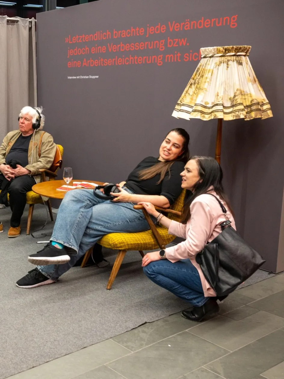 Drei Besucherinnen und Besucher der Ausstellung „Museum des Wandels" der AK Vorarlberg in der Hörstation: Eine Person sitzt mit Kopfhörer auf einem gelben Sessel und schaut auf den Bildschirm, während zwei weitere Personen daneben sitzen bzw. hocken und miteinander sprechen. Im Hintergrund ist an der grauen Wand ein Zitat aus dem Interview mit Christian Stuppner zu lesen: „Letztendlich brachte jede Veränderung jedoch eine Verbesserung bzw. eine Arbeitserleichterung mit sich."