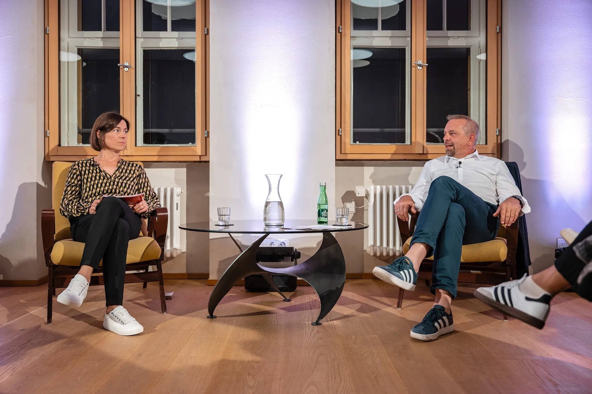 Moderatorin Carmen Jurkovic-Burtscher im Gespräch mit Oliver Loacker. Beide sitzen auf einem Sessel. In der Mitte ein Glastisch mit Getränken.