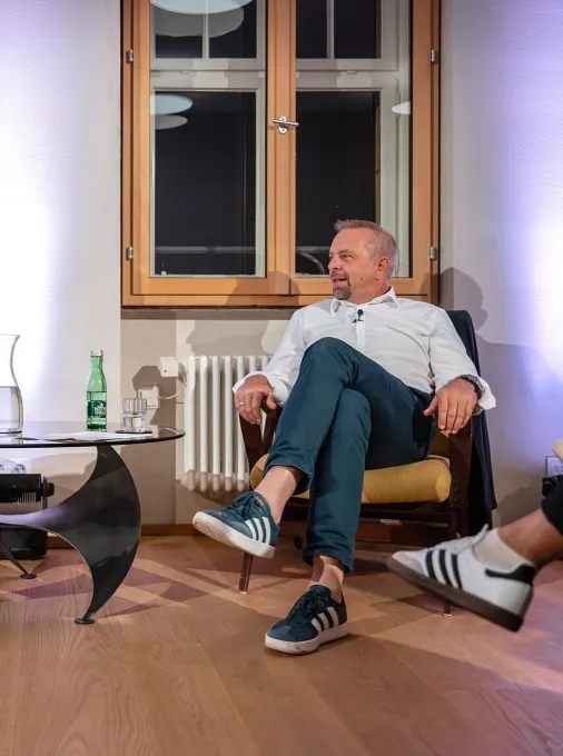 Moderatorin Carmen Jurkovic-Burtscher im Gespräch mit Oliver Loacker. Beide sitzen auf einem Sessel. In der Mitte ein Glastisch mit Getränken.