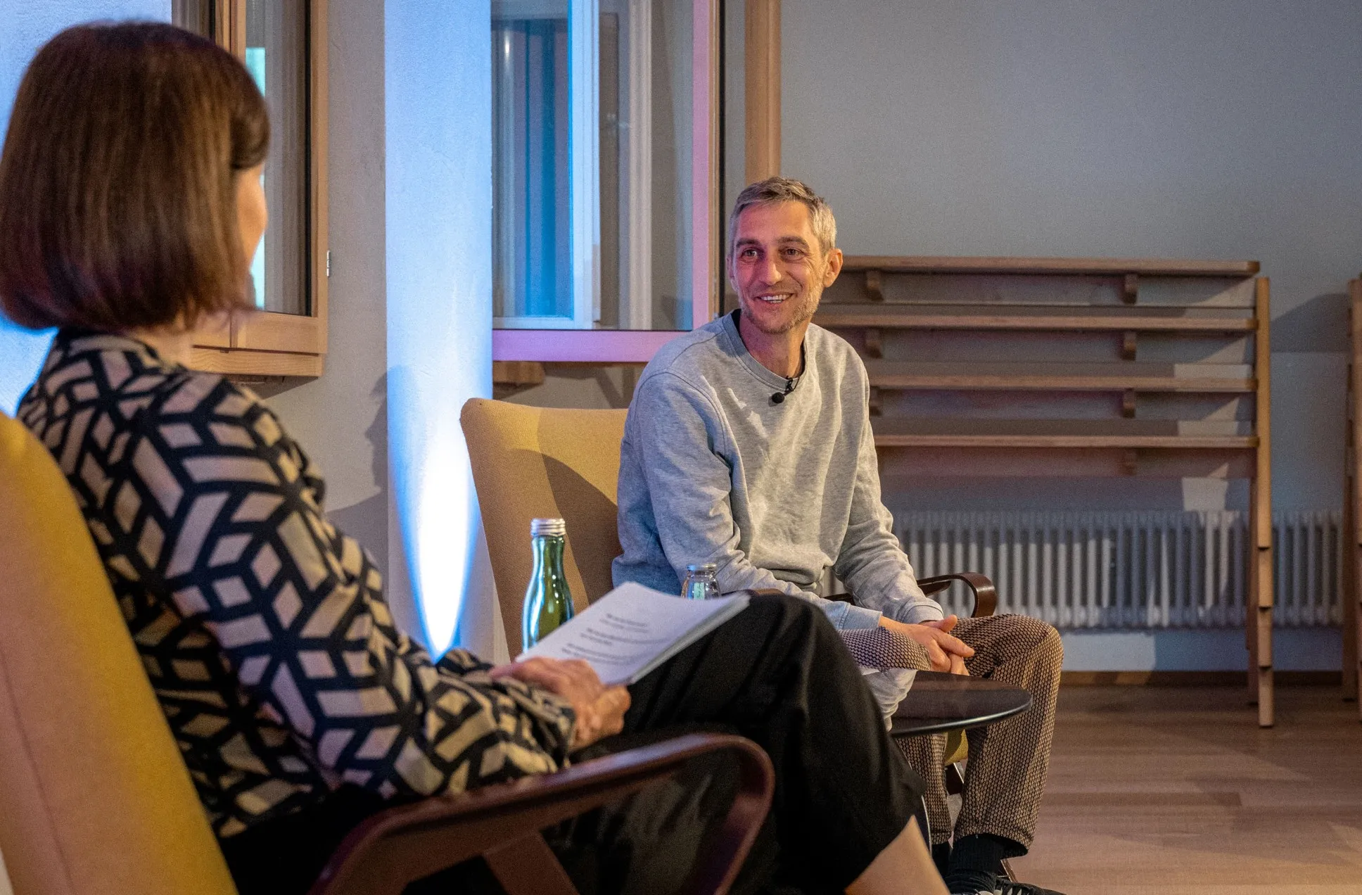 Philipp Böhler (MC Pille One) sitzt bei einer Veranstaltung der ArbeitsLebensGeschichten in der Schaffarei der AK Vorarlberg in Feldkirch auf einem Stuhl und spricht mit einer Moderatorin. Beide sind einander zugewandt, zwischen ihnen steht ein kleiner Tisch mit Wasserflaschen. Die Atmosphäre wirkt ruhig und aufmerksam, der Raum ist modern und warm beleuchtet.