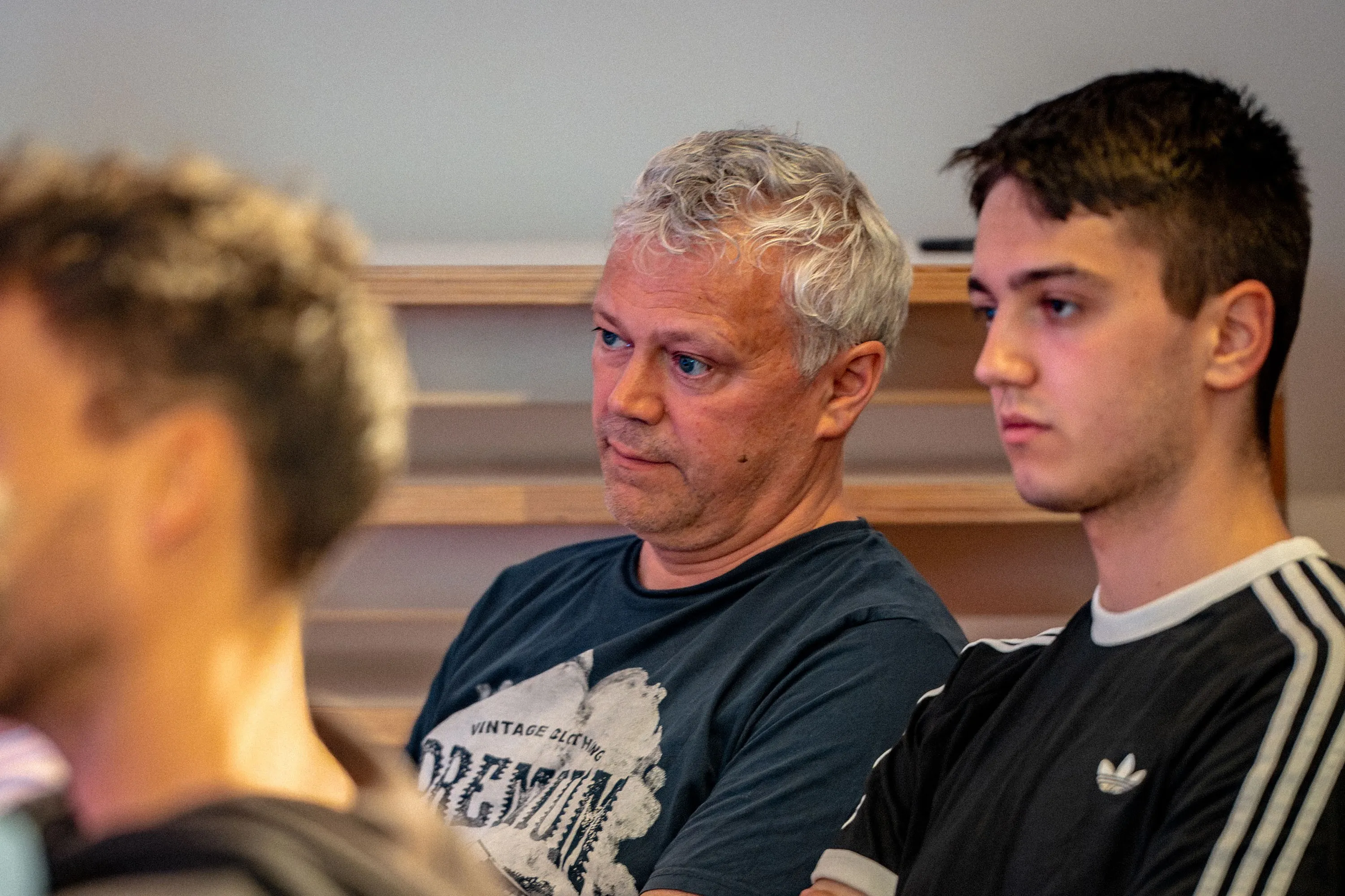 Zwei Männer sitzen nebeneinander im Publikum der Veranstaltung „ArbeitsLebensGeschichten" in der Schaffarei der Arbeiterkammer Vorarlberg in Feldkirch und verfolgen aufmerksam das Geschehen. Links ein älterer Mann mit weißem, lockigem Haar und blauen Augen im dunkelblauen Print-T-Shirt, der nachdenklich zur Seite blickt. Rechts ein jüngerer Mann mit kurzem dunklen Haar im schwarzen Adidas-T-Shirt. Im Hintergrund sind hölzerne Sitzstufen zu erkennen.