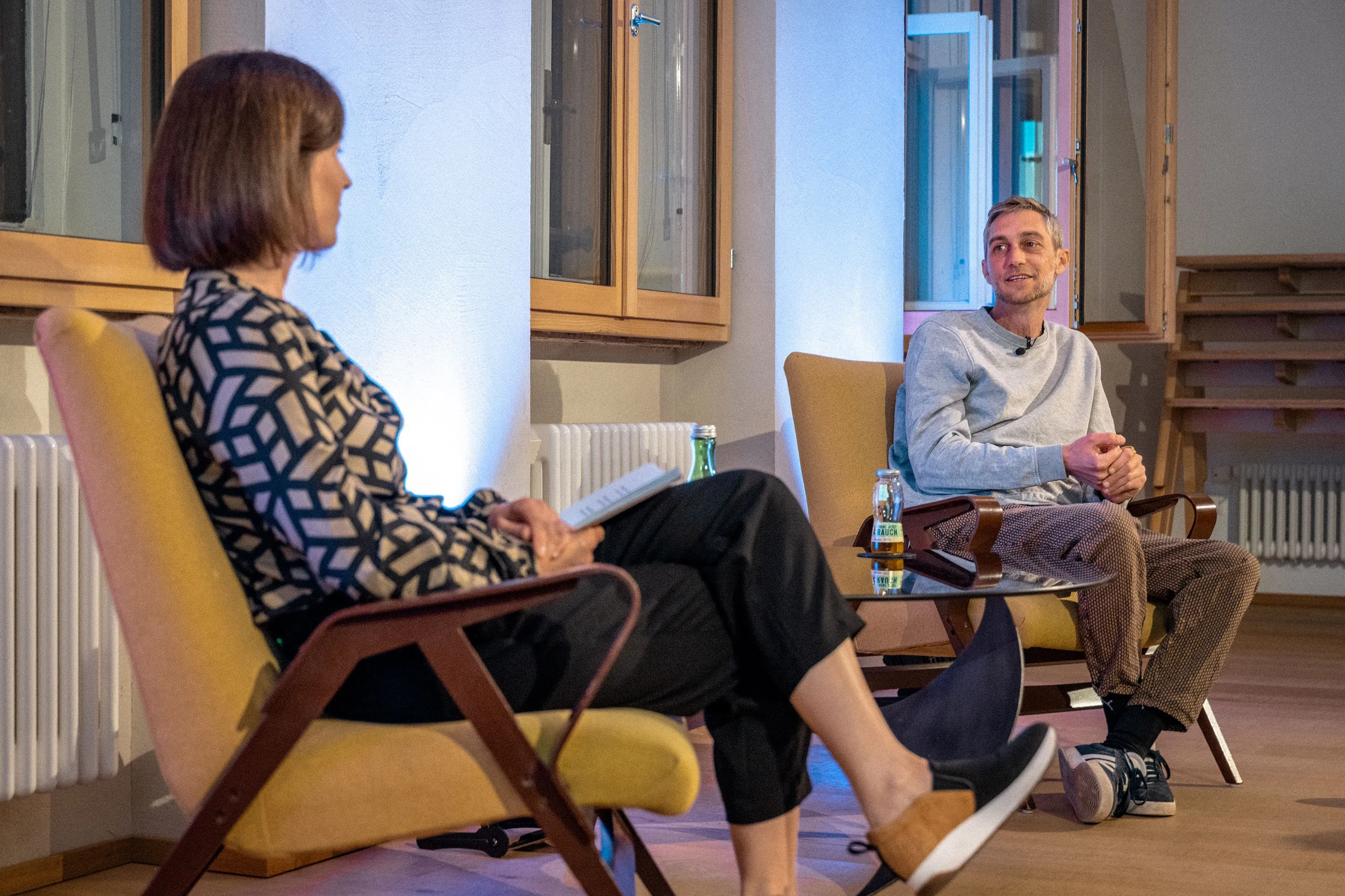 Philipp Böhler (MC Pille One) sitzt bei einer Veranstaltung der ArbeitsLebensGeschichten in der Schaffarei der AK Vorarlberg in Feldkirch auf einem Stuhl und spricht mit einer Moderatorin. Beide sind einander zugewandt, zwischen ihnen steht ein kleiner Tisch mit Wasserflaschen. Die Atmosphäre wirkt ruhig und aufmerksam, der Raum ist modern und warm beleuchtet.