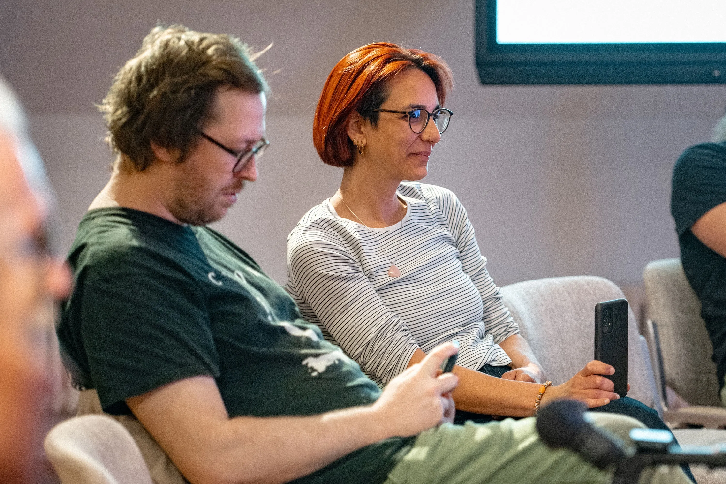 Zwei Personen sitzen nebeneinander im Publikum der Veranstaltung „ArbeitsLebensGeschichten" in der Schaffarei der Arbeiterkammer Vorarlberg in Feldkirch. Links ein junger Mann mit zerzaustem Haar und Brille im grünen T-Shirt, der auf sein Smartphone schaut. Rechts eine Frau mit kurzem roten Haar, runder Brille und gestreiftem weißen Langarmshirt, die aufmerksam nach vorne blickt und ein Smartphone in der Hand hält. Im Vordergrund ist ein Mikrofon zu erkennen, im Hintergrund eine Leinwand.