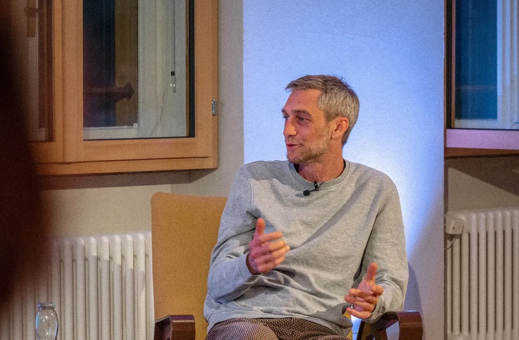 Philipp Böhler (MC Pille One) sitzt bei einer Veranstaltung der ArbeitsLebensGeschichten in der Schaffarei der AK Vorarlberg in Feldkirch auf einem Stuhl und spricht mit einer Moderatorin. Beide sind einander zugewandt, zwischen ihnen steht ein kleiner Tisch mit Wasserflaschen. Die Atmosphäre wirkt ruhig und aufmerksam, der Raum ist modern und warm beleuchtet.