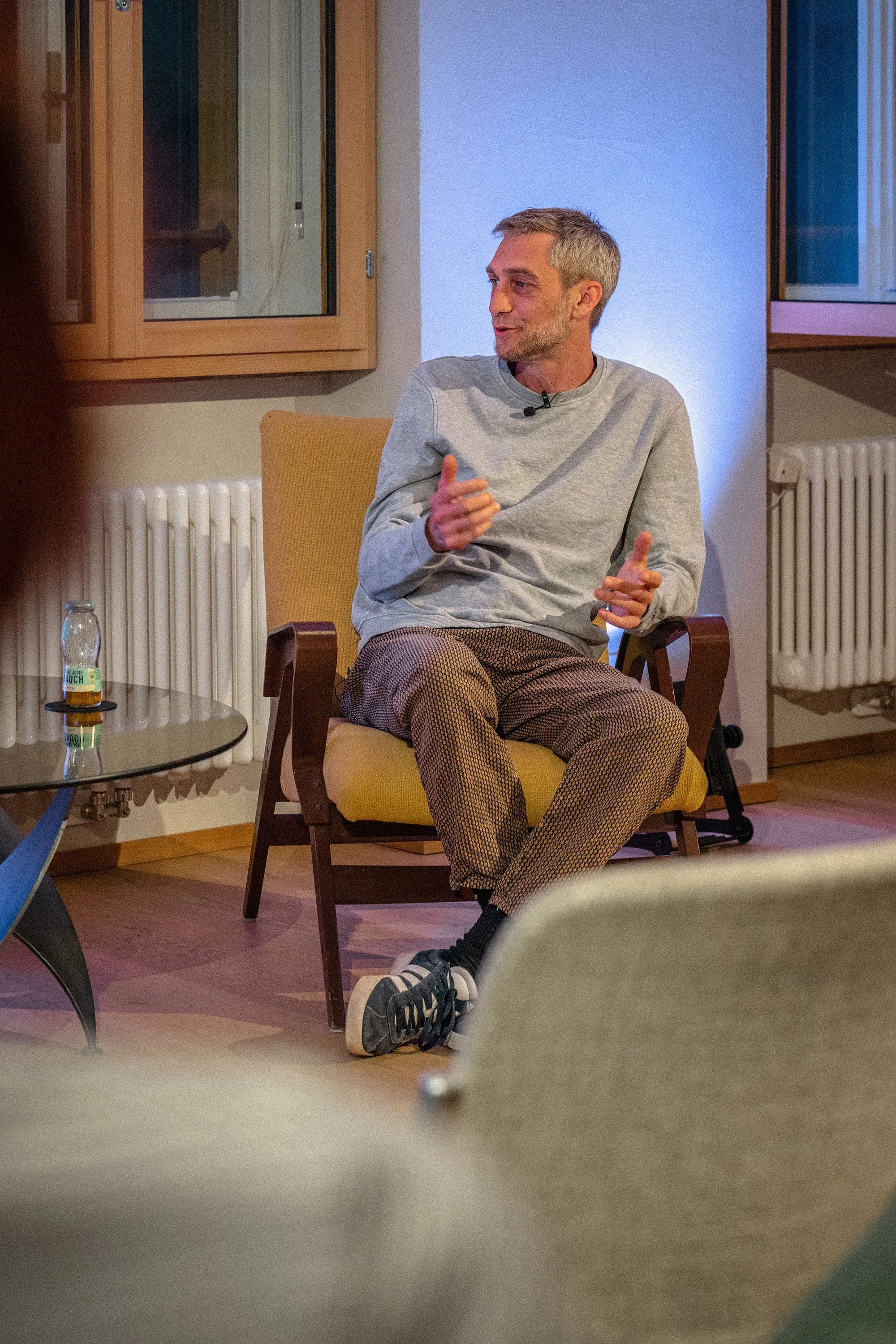 Philipp Böhler (MC Pille One) sitzt bei einer Veranstaltung der ArbeitsLebensGeschichten in der Schaffarei der AK Vorarlberg in Feldkirch auf einem Stuhl und spricht mit einer Moderatorin. Beide sind einander zugewandt, zwischen ihnen steht ein kleiner Tisch mit Wasserflaschen. Die Atmosphäre wirkt ruhig und aufmerksam, der Raum ist modern und warm beleuchtet.