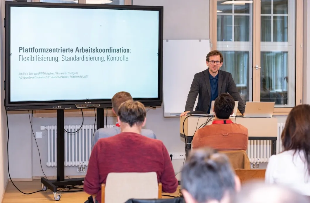 Vortragender mit Brille mit einem großem Bildschirm und Seminarbesucher im Vordergrund