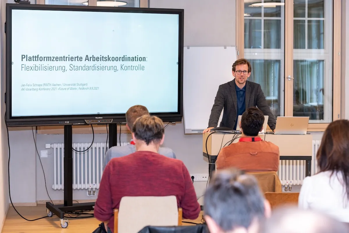 Vortragender mit Brille mit einem großem Bildschirm und Seminarbesucher im Vordergrund