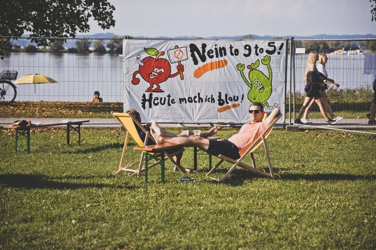 Zwei Personen im Liegestuhl und im Hintergrund ein Plakat und ein See