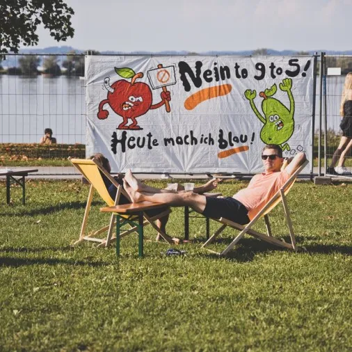 Zwei Personen im Liegestuhl und im Hintergrund ein Plakat und ein See