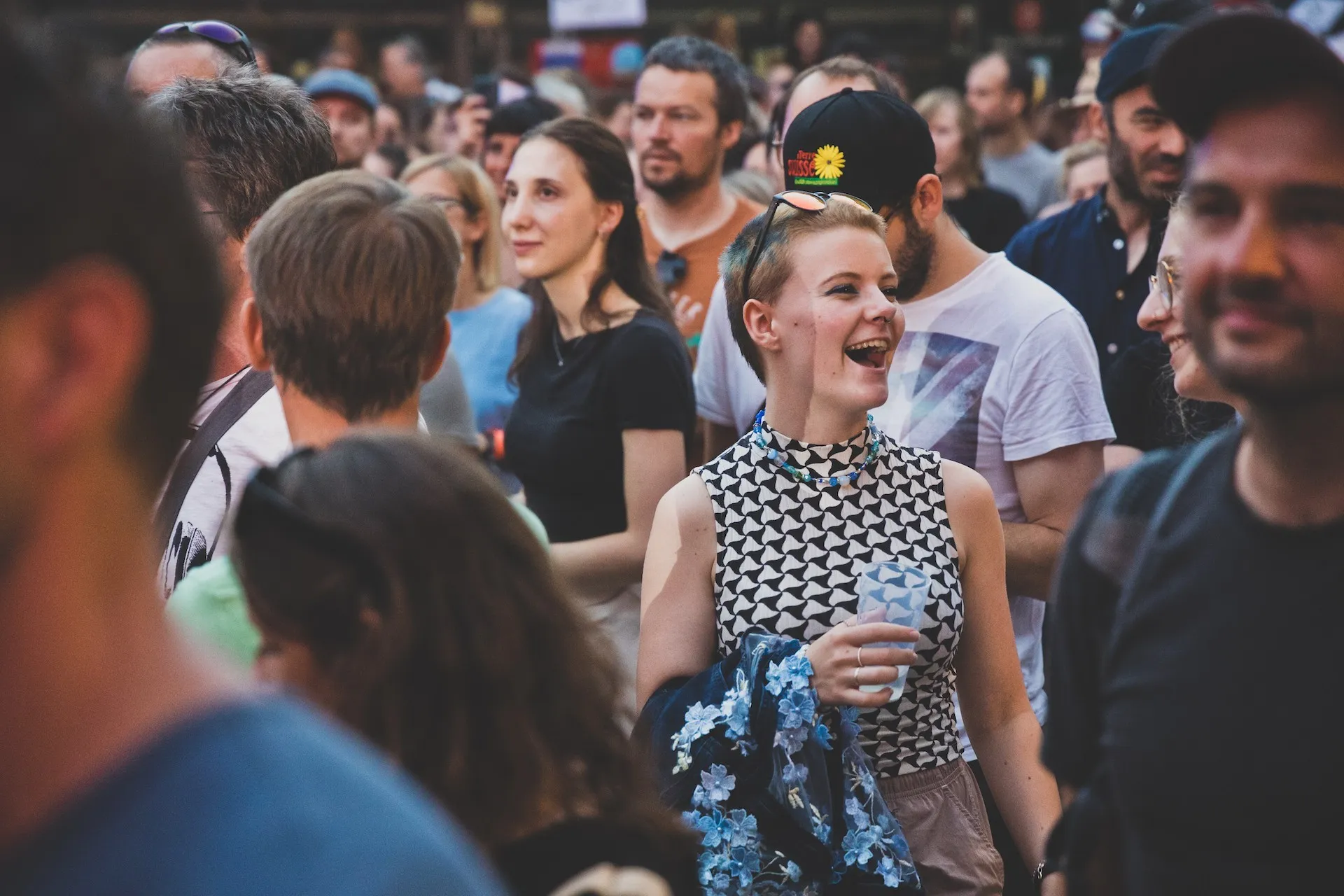 Frau steht im Mittelpunkt lachend mit Getränk in der Hand und Menschenmengen im Hintergrund