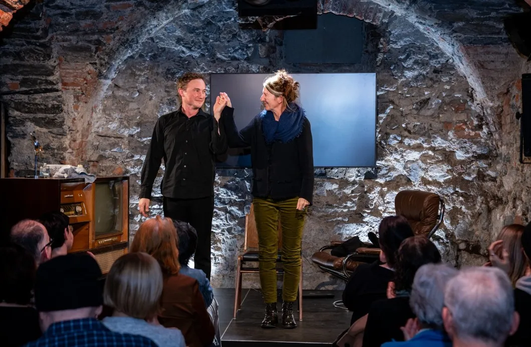 Schauspieler David Kopp und Regisseurin Daniela Egger verbeugen sich gemeinsam beim Schlussapplaus auf der Bühne des Schaffarei Clubs in Feldkirch nach der Vorstellung von „Move fast and break things". Kopp trägt ein schwarzes Hemd und schwarze Hose, Egger einen dunklen Pullover mit blauem Schal und olivgrüner Hose. Sie halten sich an den Händen und lächeln einander an. Im Hintergrund ist die beleuchtete Natursteinwand mit einem dunklen Bildschirm zu sehen, links ein alter Röhrenfernseher als Bühnendekoration. Zu beiden Seiten sitzt das applaudierende Publikum.