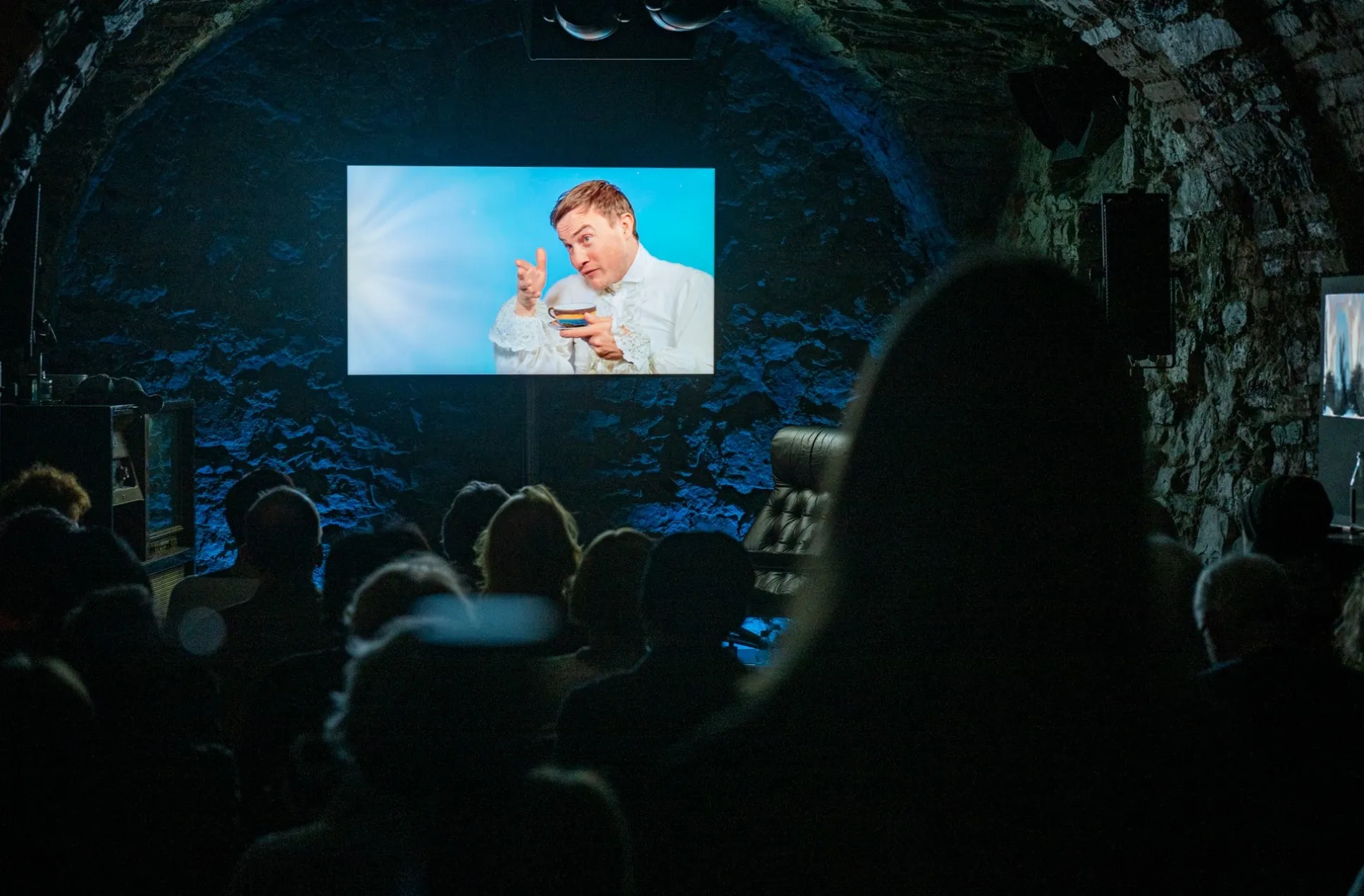 Blick in den gewölbten Zuschauerraum des Schaffarei Clubs in Feldkirch während einer Vorstellung von „Move fast and break things" von Daniela Egger. Das Publikum sitzt im Dunkeln und schaut auf eine Leinwand, auf der ein Videoeinspieler mit Schauspieler David Kopp zu sehen ist. Er trägt ein weißes, historisch anmutendes Kostüm mit Rüschen und hält eine Tasse in der Hand, während er mit aufgerissenen Augen und erhobenem Zeigefinger in die Kamera schaut. Der Raum ist aus Naturstein gemauert und in blaues Licht getaucht.