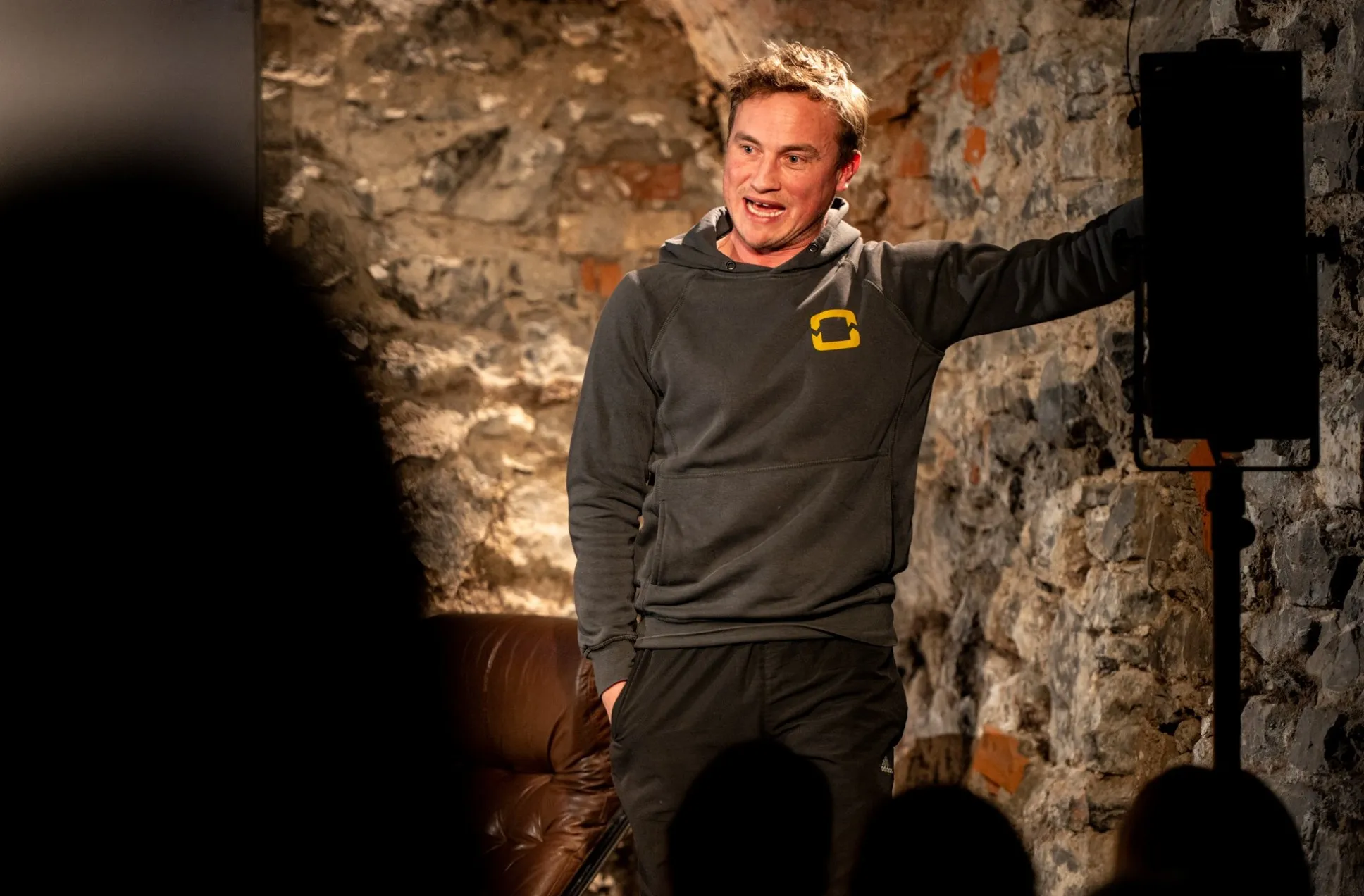 Schauspieler David Kopp steht auf der Bühne des Schaffarei Clubs in Feldkirch und spielt in dem Theaterstück „Move fast and break things" von Daniela Egger. Er trägt einen dunkelgrauen Hoodie mit gelbem Logo und schwarze Jogginghose und lehnt mit dem rechten Arm an der beleuchteten Natursteinwand. Sein Mund ist leicht geöffnet, als ob er gerade spricht. Im Vordergrund sind die Silhouetten von Zuschauern zu sehen.