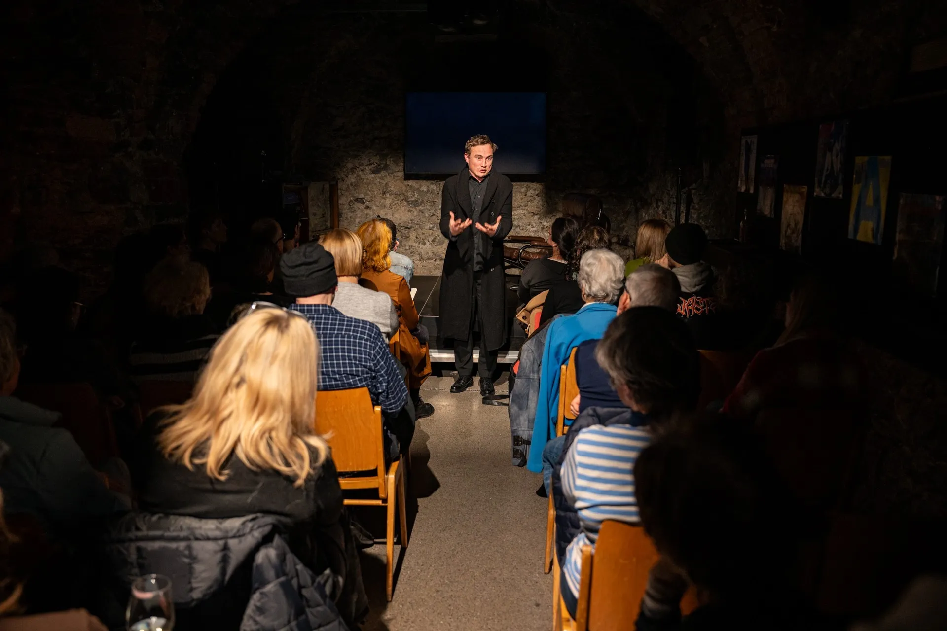 Blick in den voll besetzten Zuschauerraum des Schaffarei Clubs in Feldkirch während einer Vorstellung von „Move fast and break things" von Daniela Egger. Das Publikum sitzt auf Holzstühlen in zwei Reihen zu beiden Seiten eines schmalen Mittelgangs. Am Ende des Gangs steht Schauspieler David Kopp in einem langen schwarzen Mantel und gestikuliert mit beiden Händen. Im Hintergrund ist die beleuchtete Natursteinwand sowie ein dunkler Bildschirm unter dem Gewölbe zu sehen.